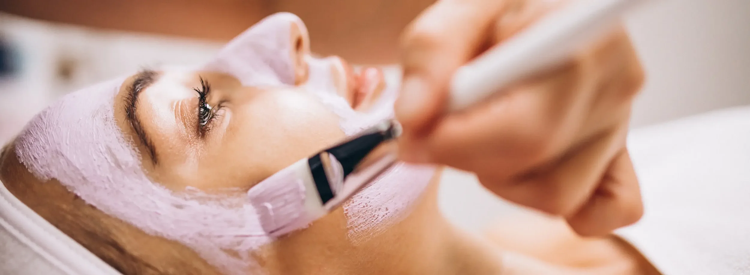 A woman lying down with her eyes closed, receiving a facial treatment with a pink facial mask.
