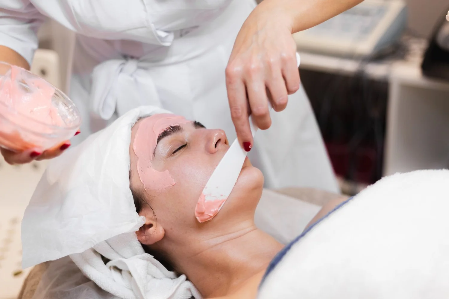 A woman is receiving a facial treatment with a pink facial mask applied on her face at a spa or clinic.