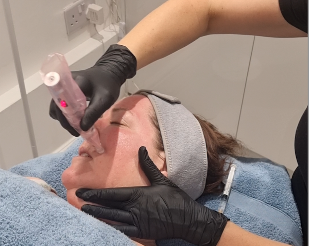 Medical professional administering a facial injection to a woman lying on a treatment bed, with a towel covering part of her body, in a clinical setting.