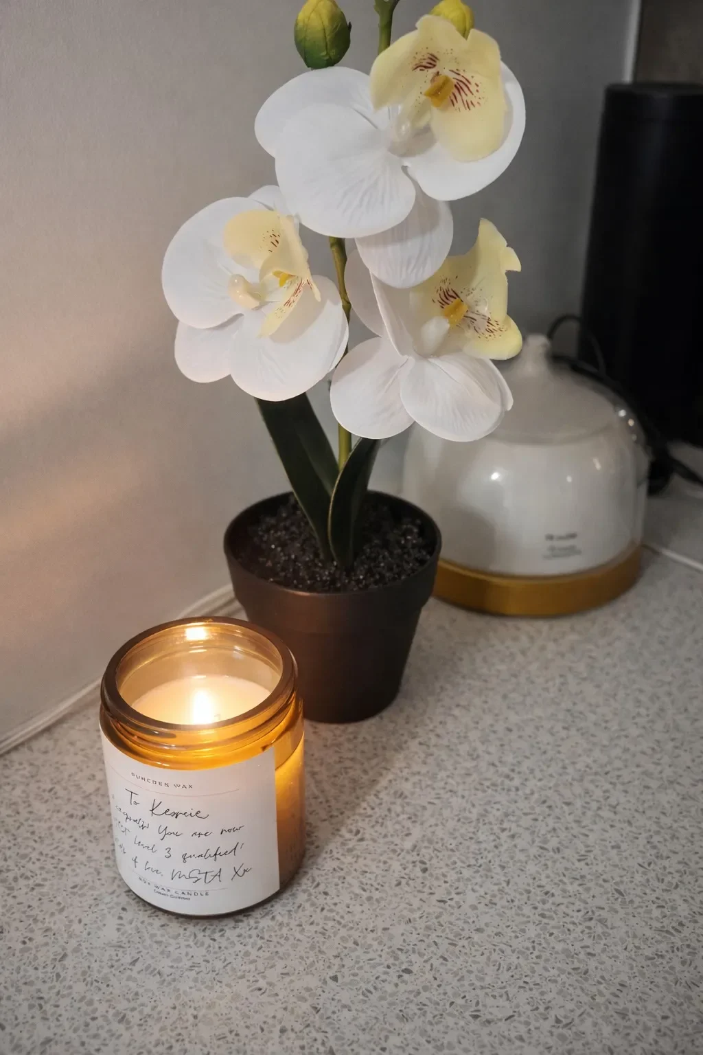 A white orchid potted plant, a lit candle in a white jar with handwritten label, and a white electric kettle on a kitchen countertop.