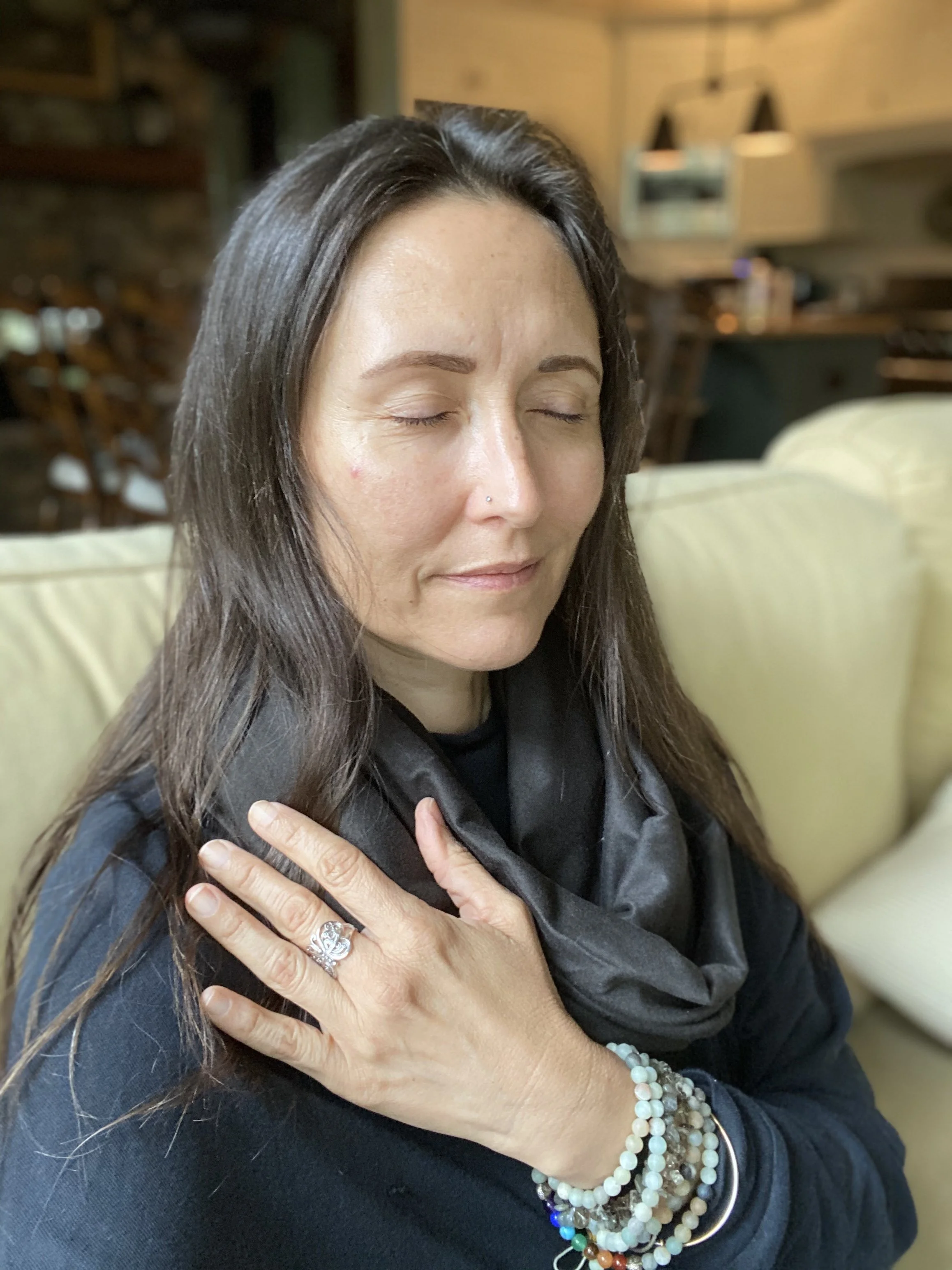 A woman with long dark hair and a nose piercing is sitting on a beige sofa with her eyes closed and hands crossed over her chest, wearing multiple bead bracelets and a large ring.