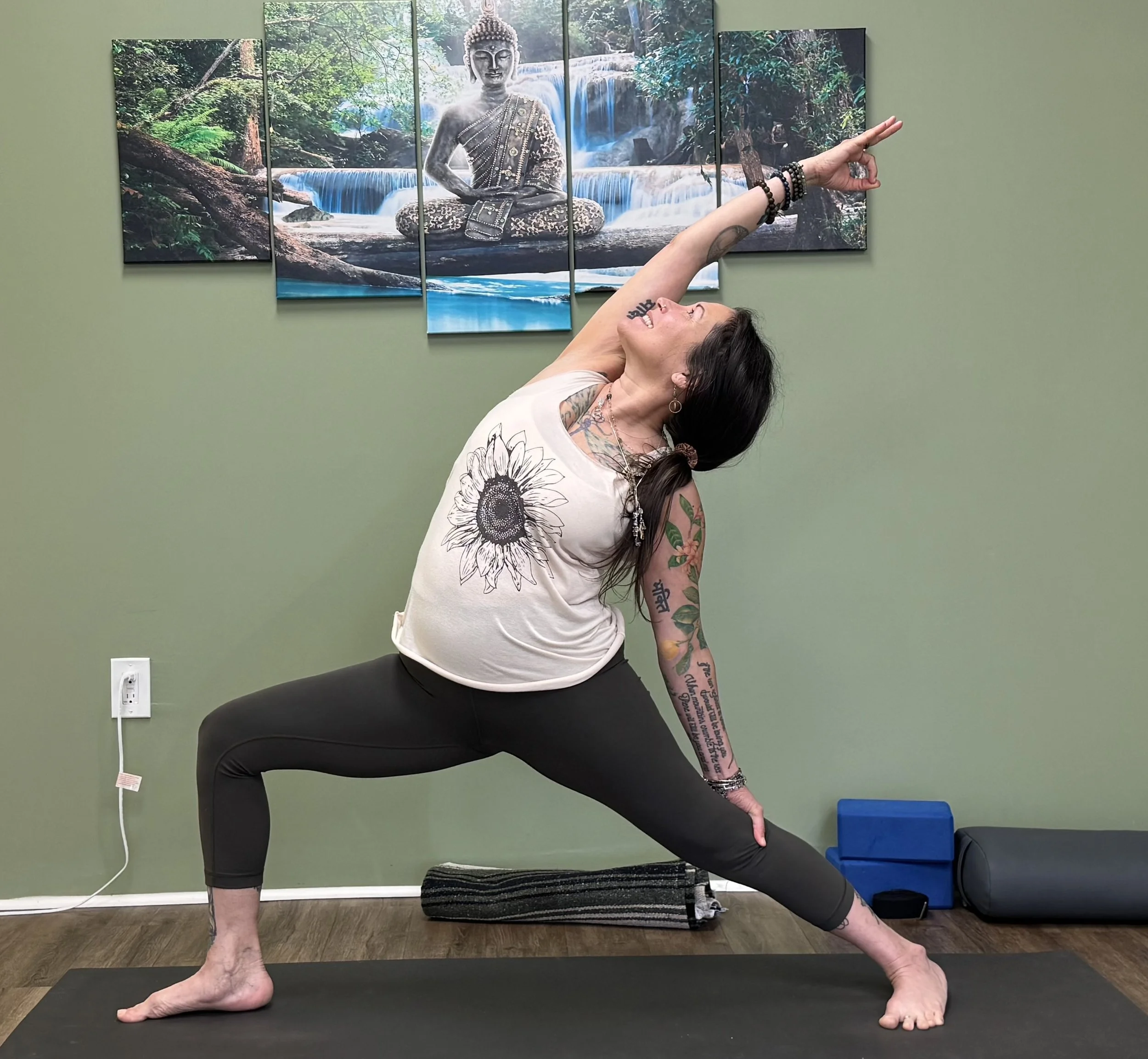 A woman with tattoos and bracelets doing a yoga pose in front of a four-panel painting of a Buddha statue in a forest with a waterfall, in a room with a green wall.
