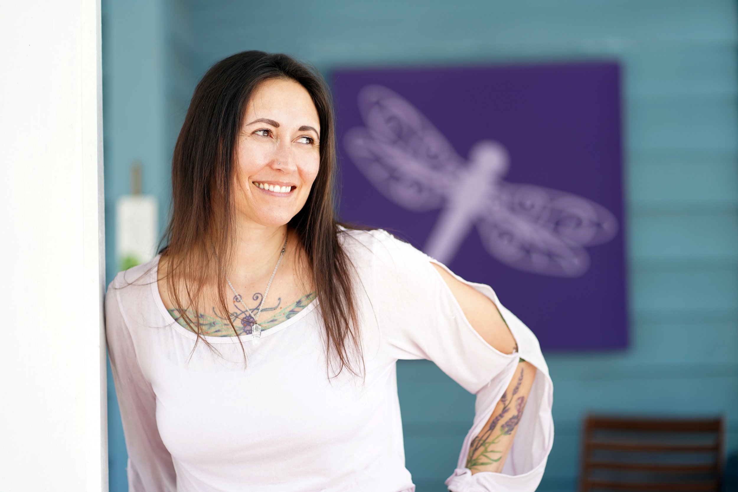 A woman with long dark hair, a nose piercing, and colorful tattoos on her chest and arms, smiling and looking to the side, standing indoors near a white wall with a blue background and a purple art piece.