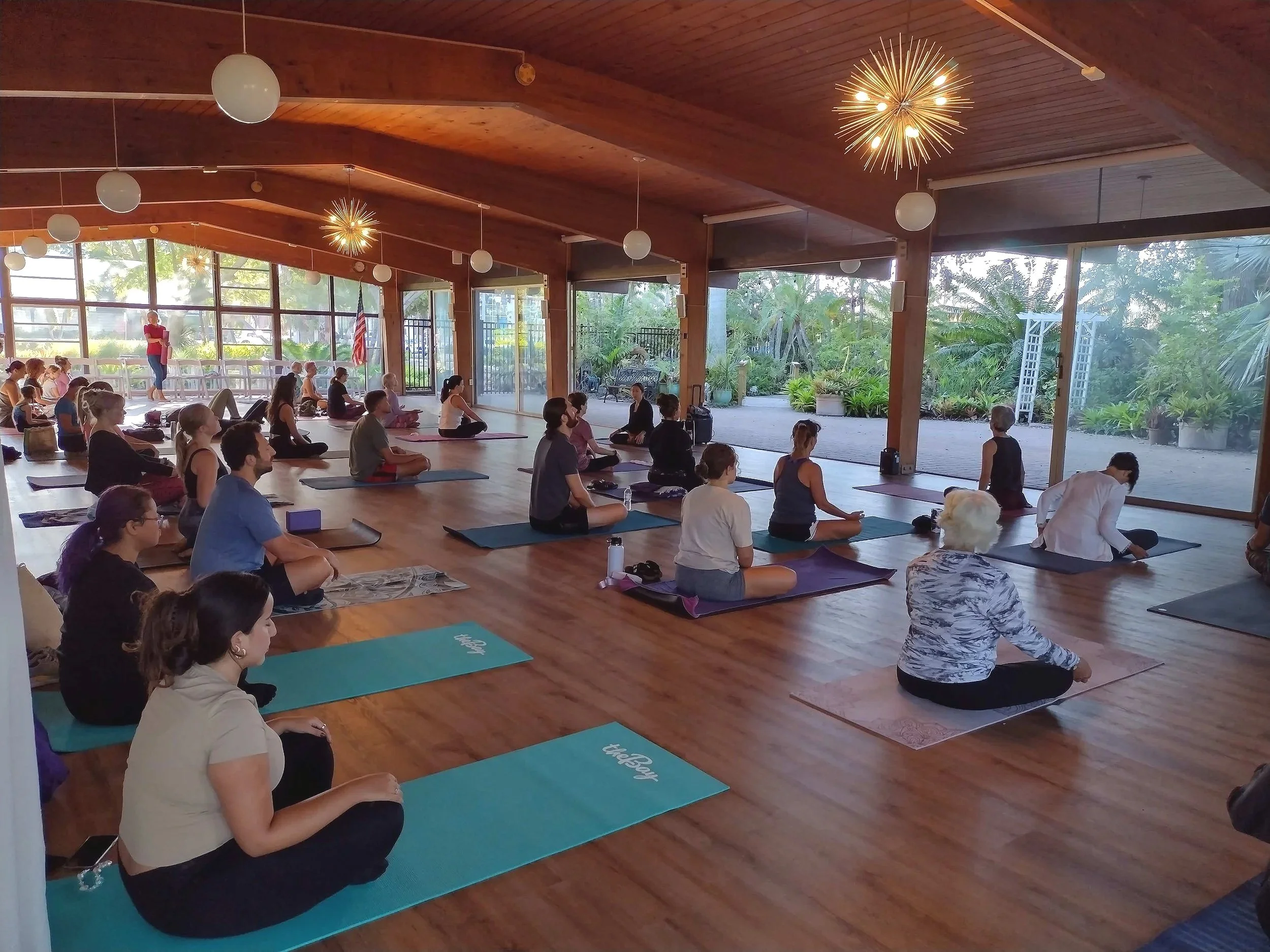 People participating in a group yoga or meditation class in a sunlit indoor space with large windows, wooden ceiling, and outdoor greenery.