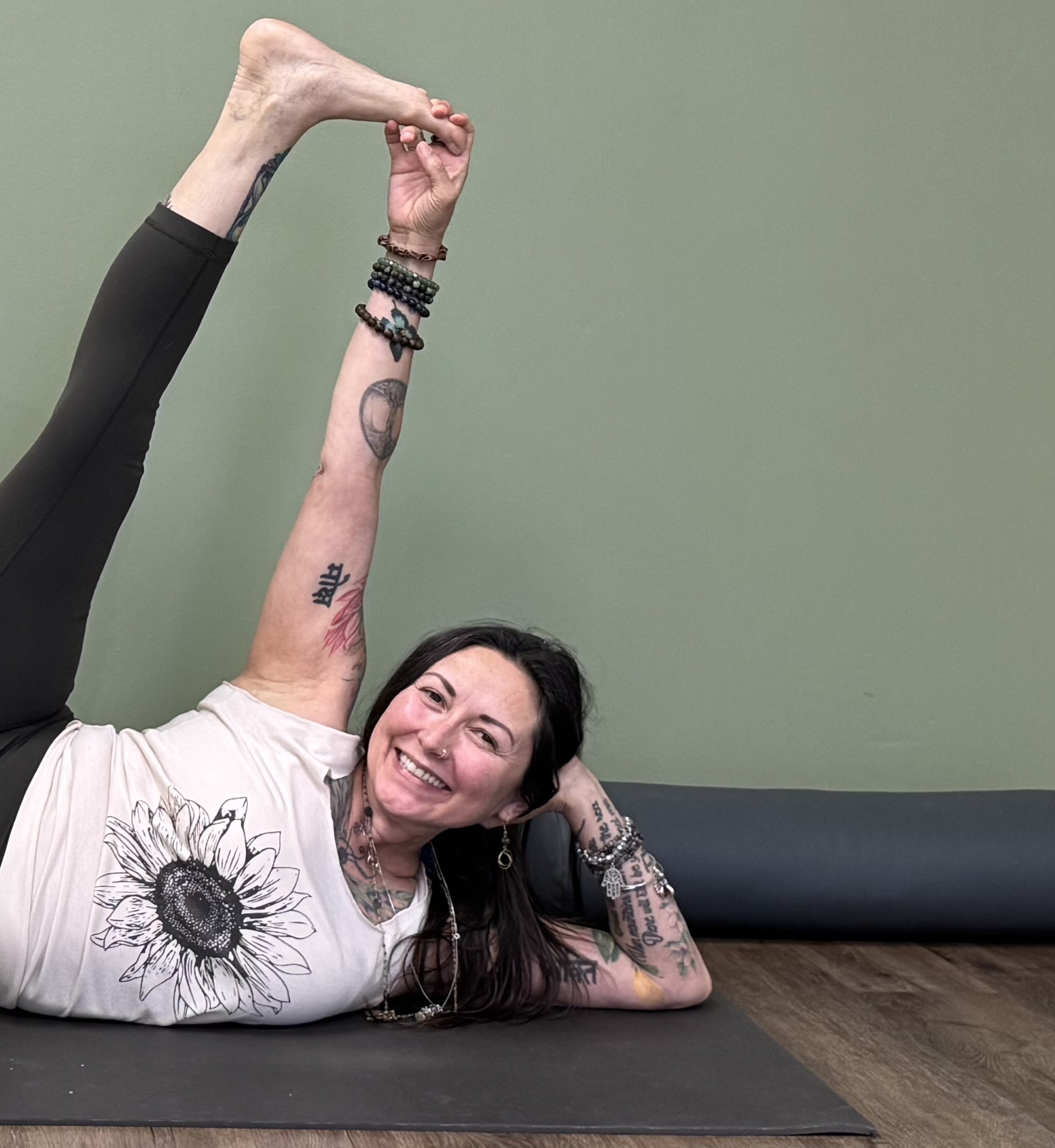 A woman with dark hair, tattoos, and a nose piercing, smiling while lying on her side on a yoga mat, her legs raised and intertwined with another person's legs, practicing yoga in a room with a plain green wall.