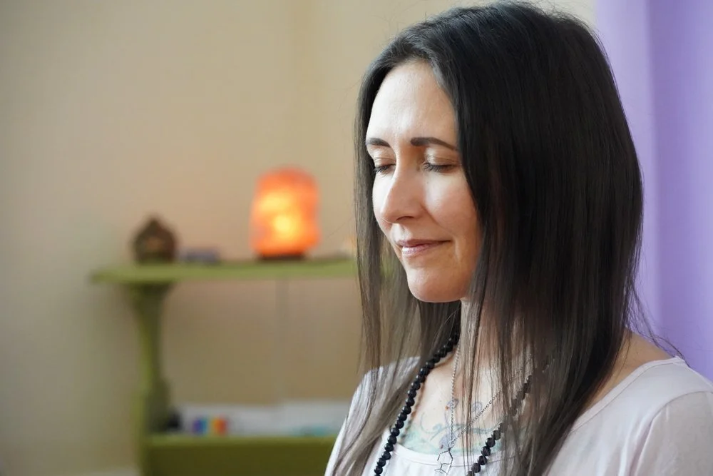 A woman with long dark hair and closed eyes, smiling softly, in a room with a green shelf and decorative lamp in the background.