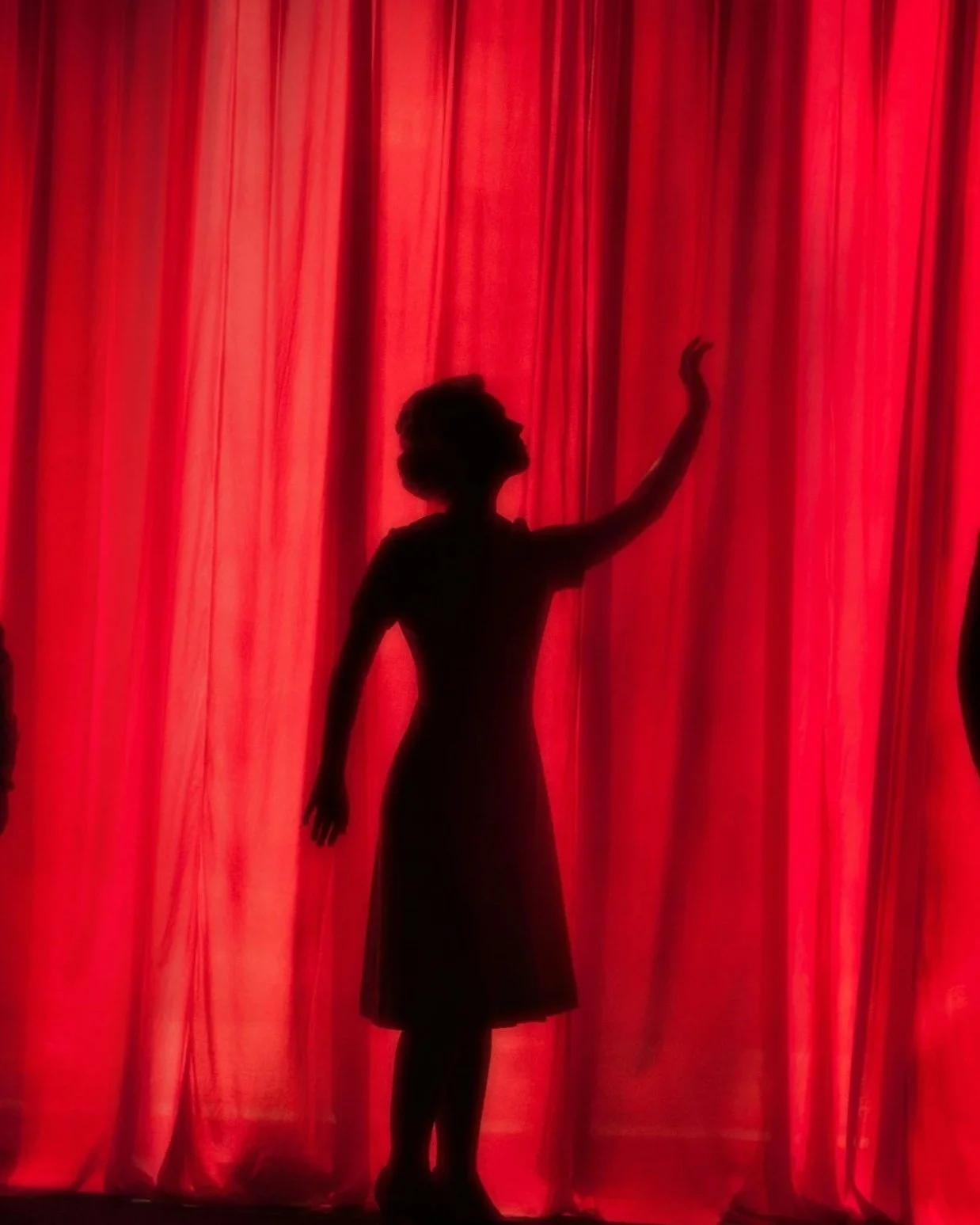 Silhouette of a woman on stage in front of red curtains, waving with her right hand.