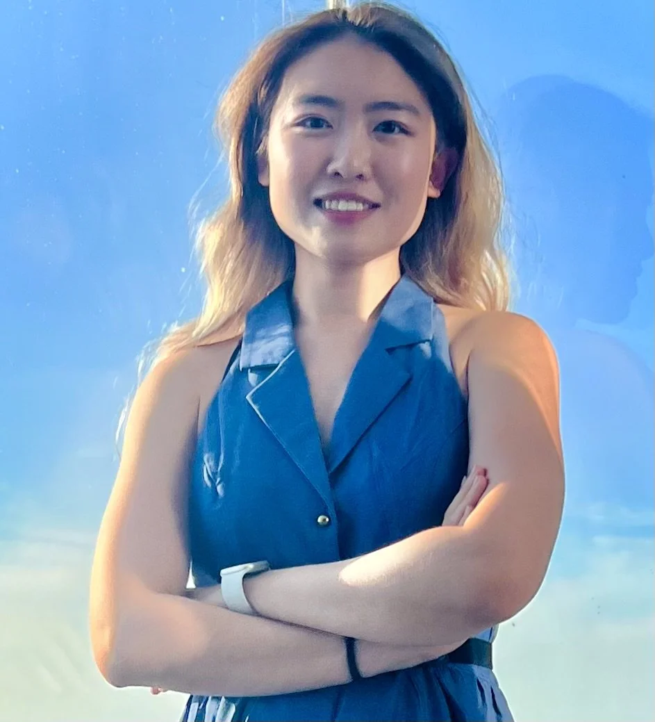 A smiling woman with long, wavy hair wearing a sleeveless blue shirt, standing with her arms crossed in front of a blue background.