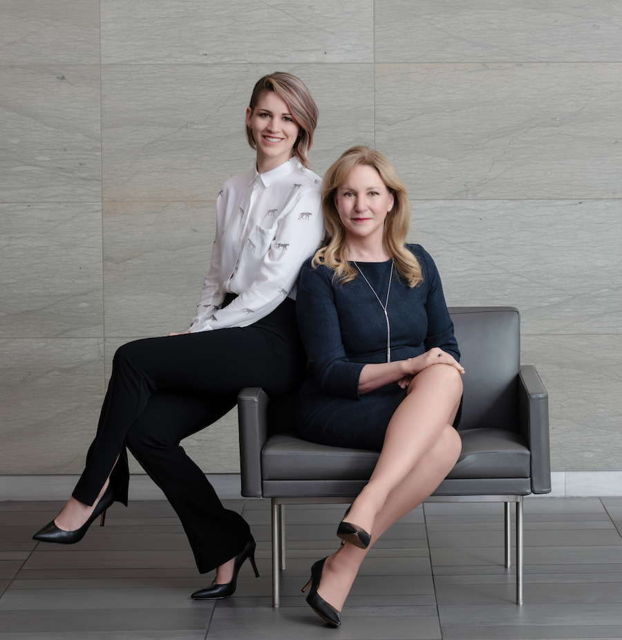 Two women in business attire pose in a modern office setting, one seated on a gray chair with crossed legs and the other standing behind her, leaning on the chair.