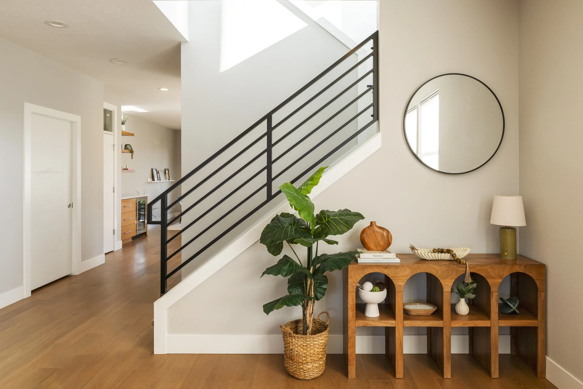 A modern living space with a staircase, a large potted plant, a wooden console table with decorative items, a round mirror, and a table lamp.