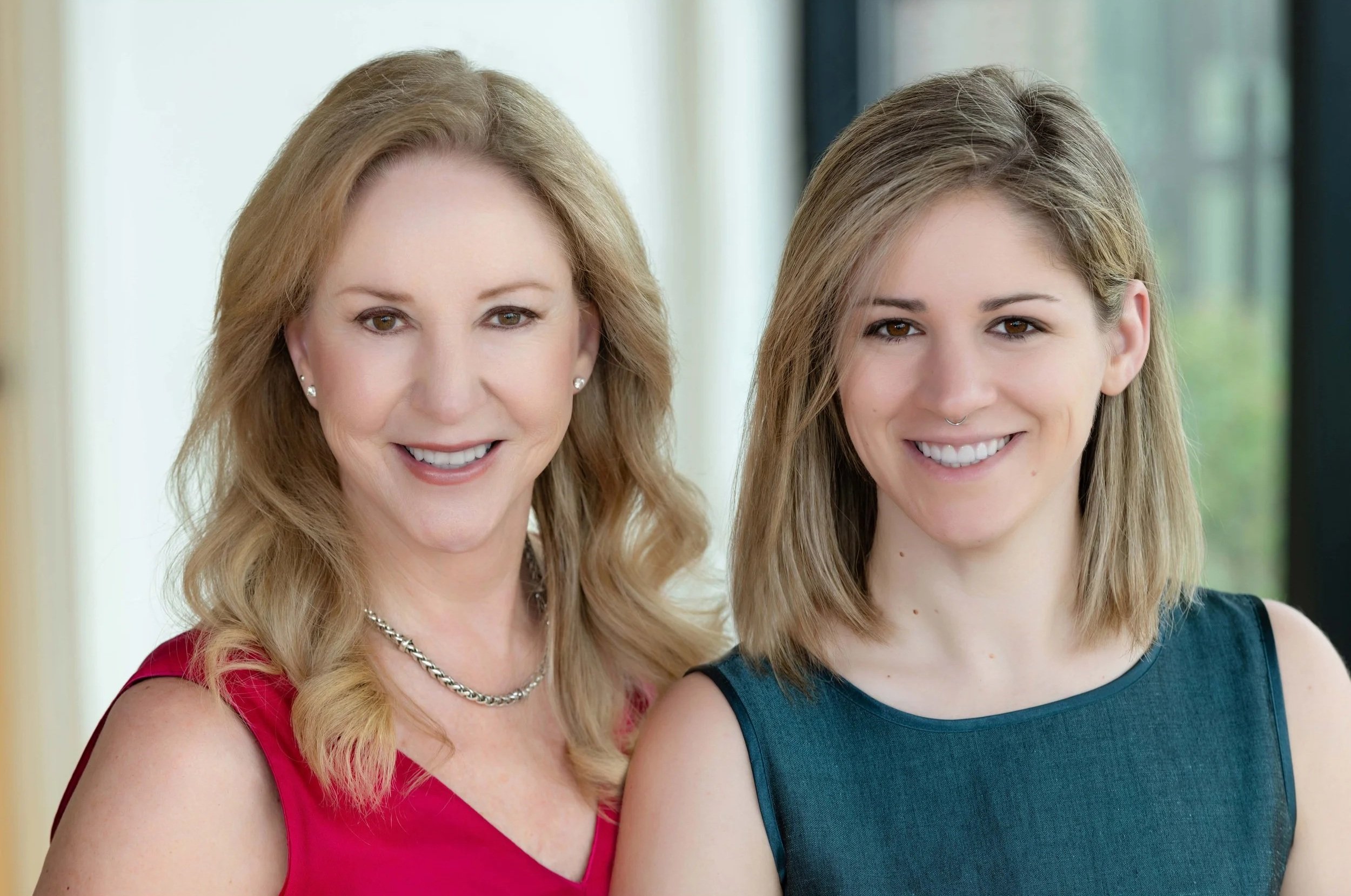 Two welcoming business women, a mother-daughter team, smiling indoors with a window in the background.