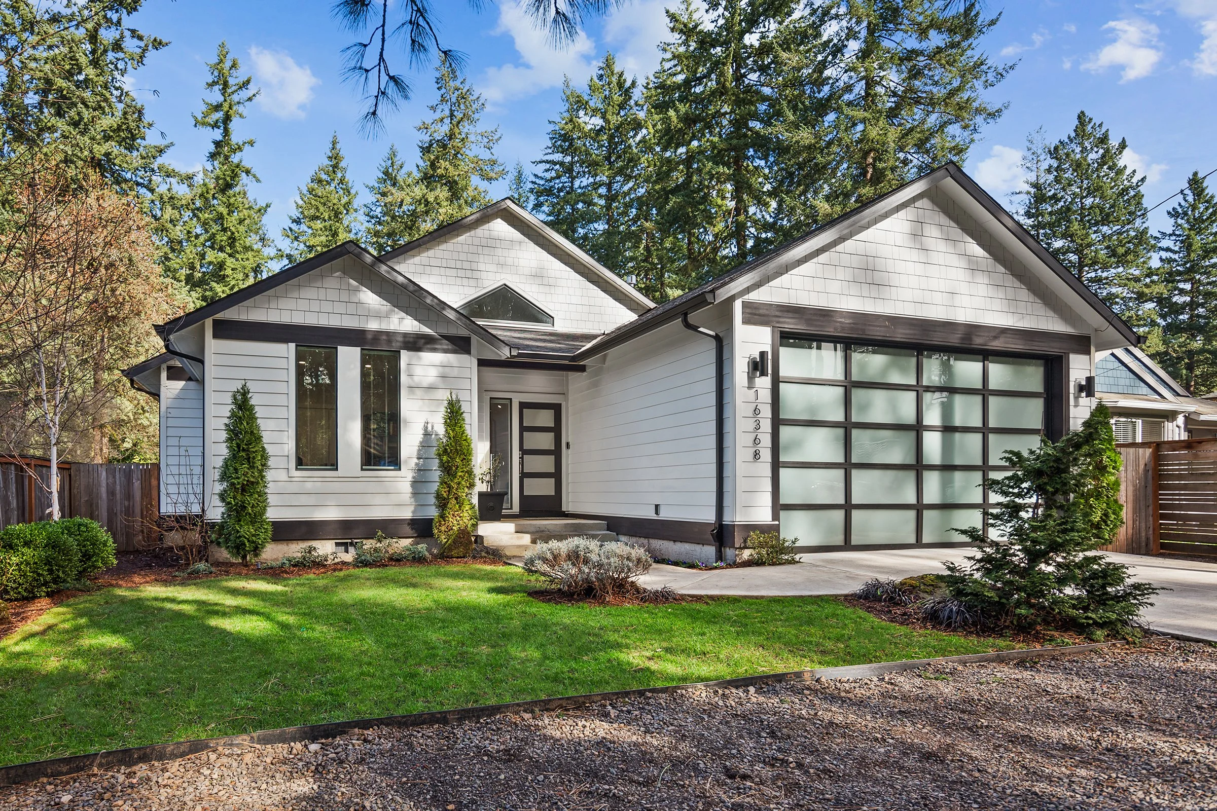 A modern house with a white exterior, large frosted glass garage door, and black trim, surrounded by a green lawn with small shrubs and tall trees in the background.