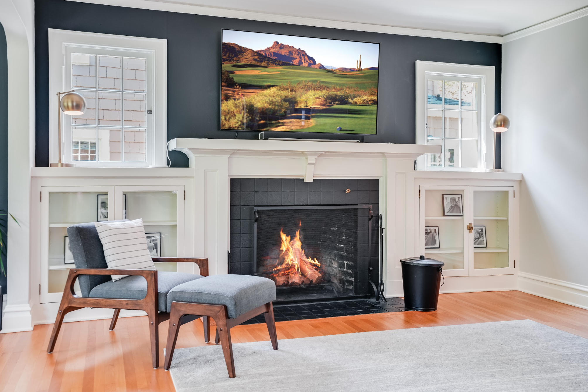 Living room with a fireplace, a television mounted above it displaying a scenic landscape, and a modern armchair with a footrest and striped pillow in front of the fireplace.