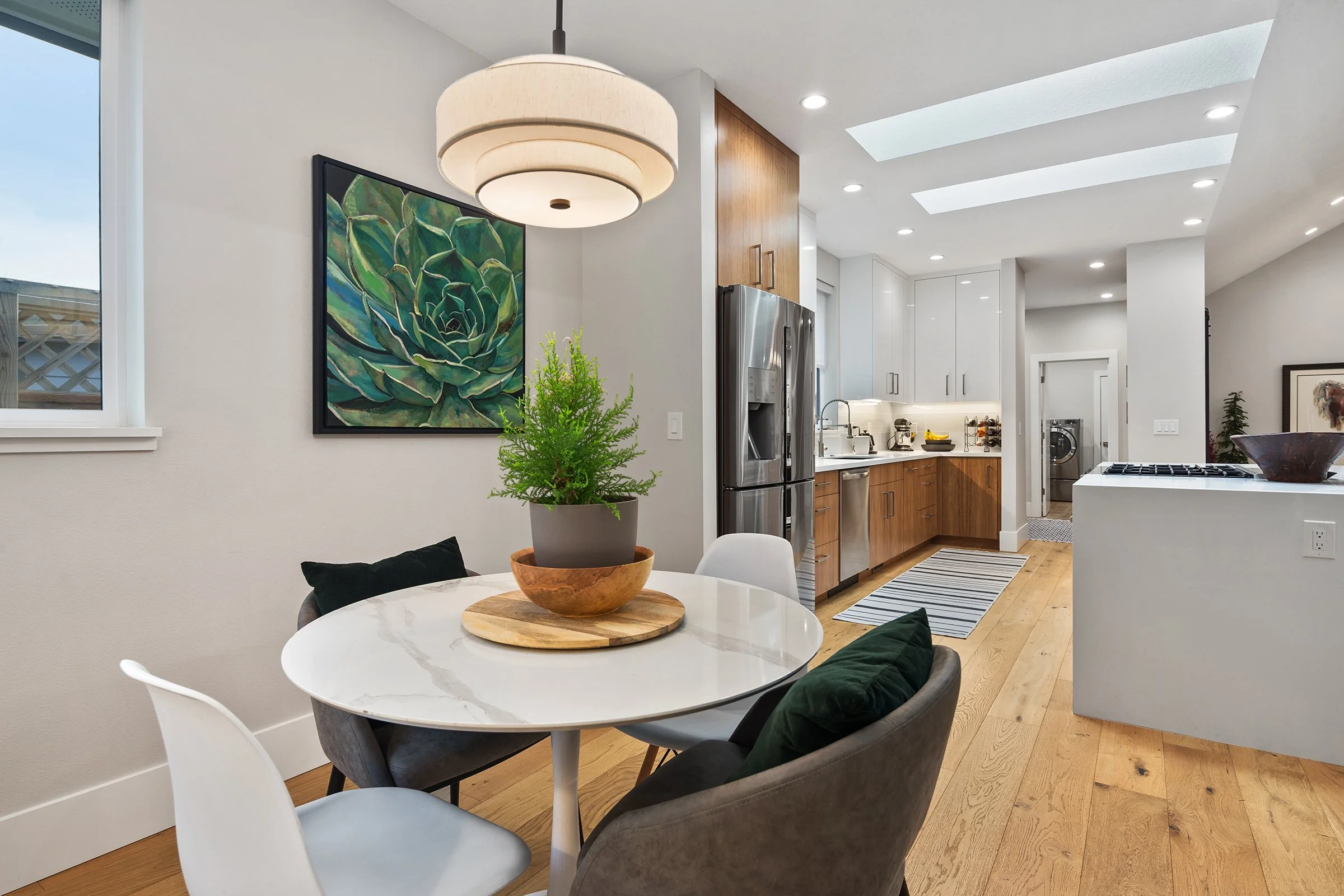 A modern kitchen and dining area with a round white table, four chairs, a potted plant, window with a view, and kitchen appliances including a stainless steel refrigerator.