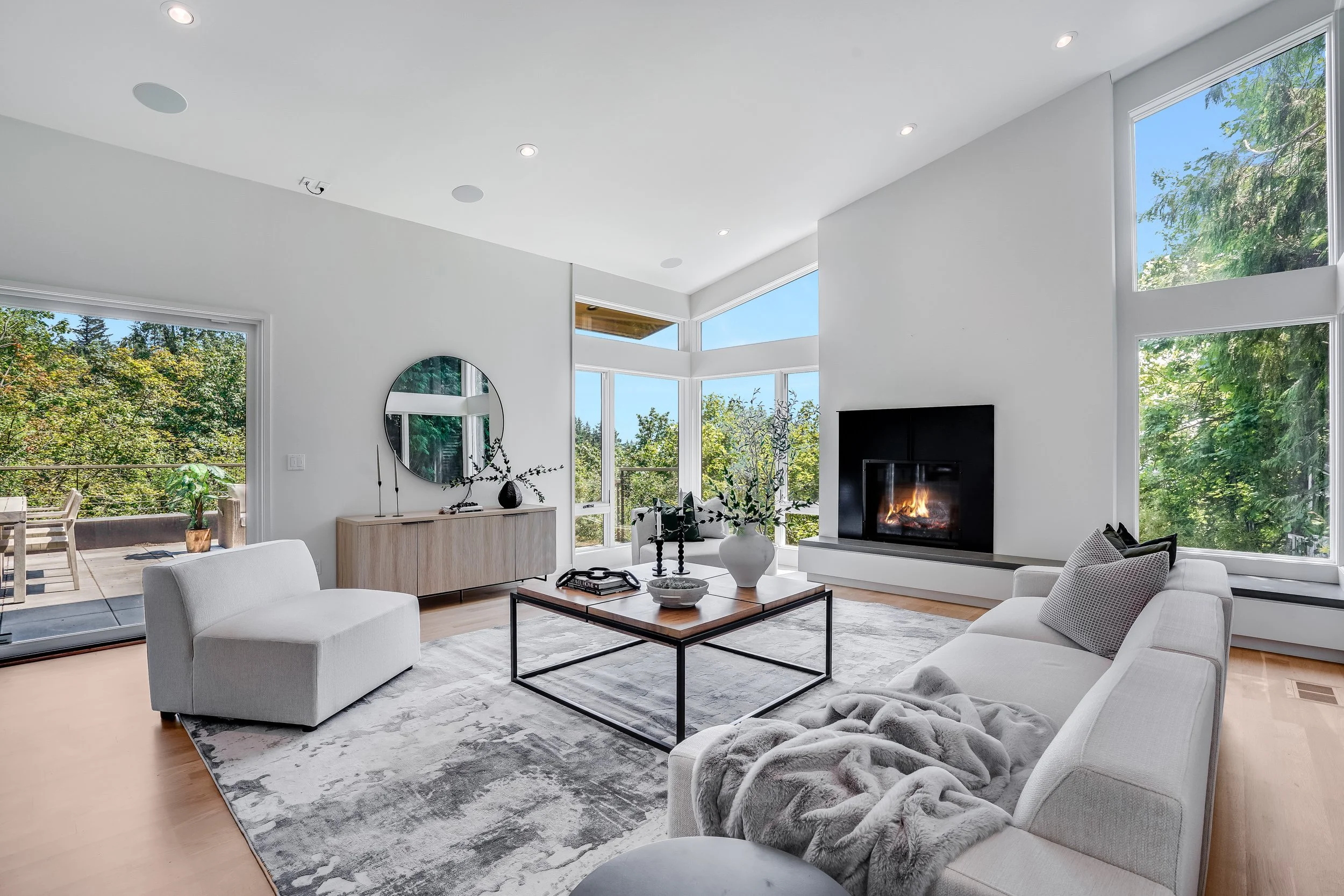 Living room with white couches, a coffee table, a fireplace, and large windows showing greenery outside.