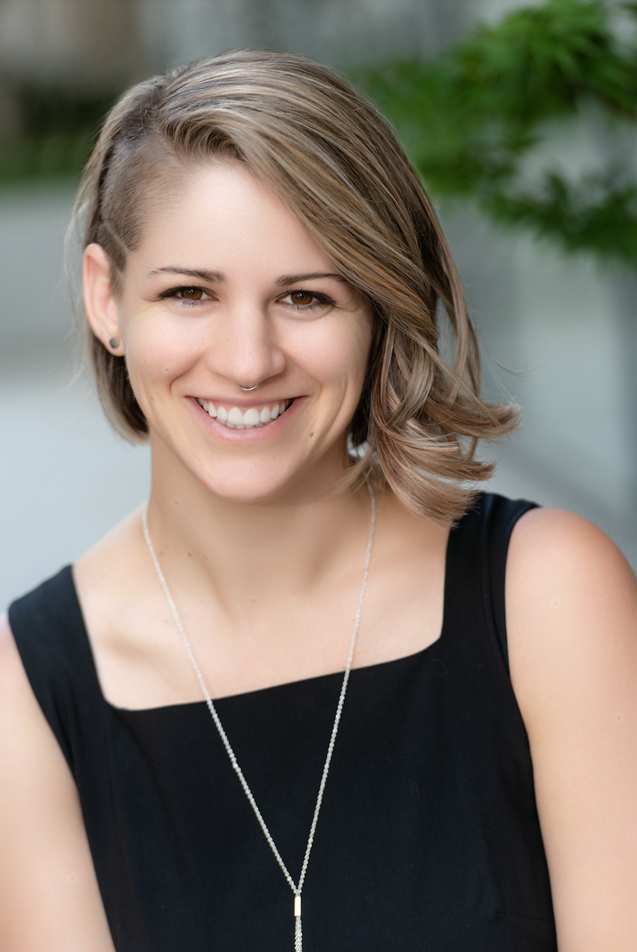 Portrait of a young woman with short styled hair, smiling, wearing a black sleeveless top and a silver necklace, outdoors with blurred green foliage in the background.