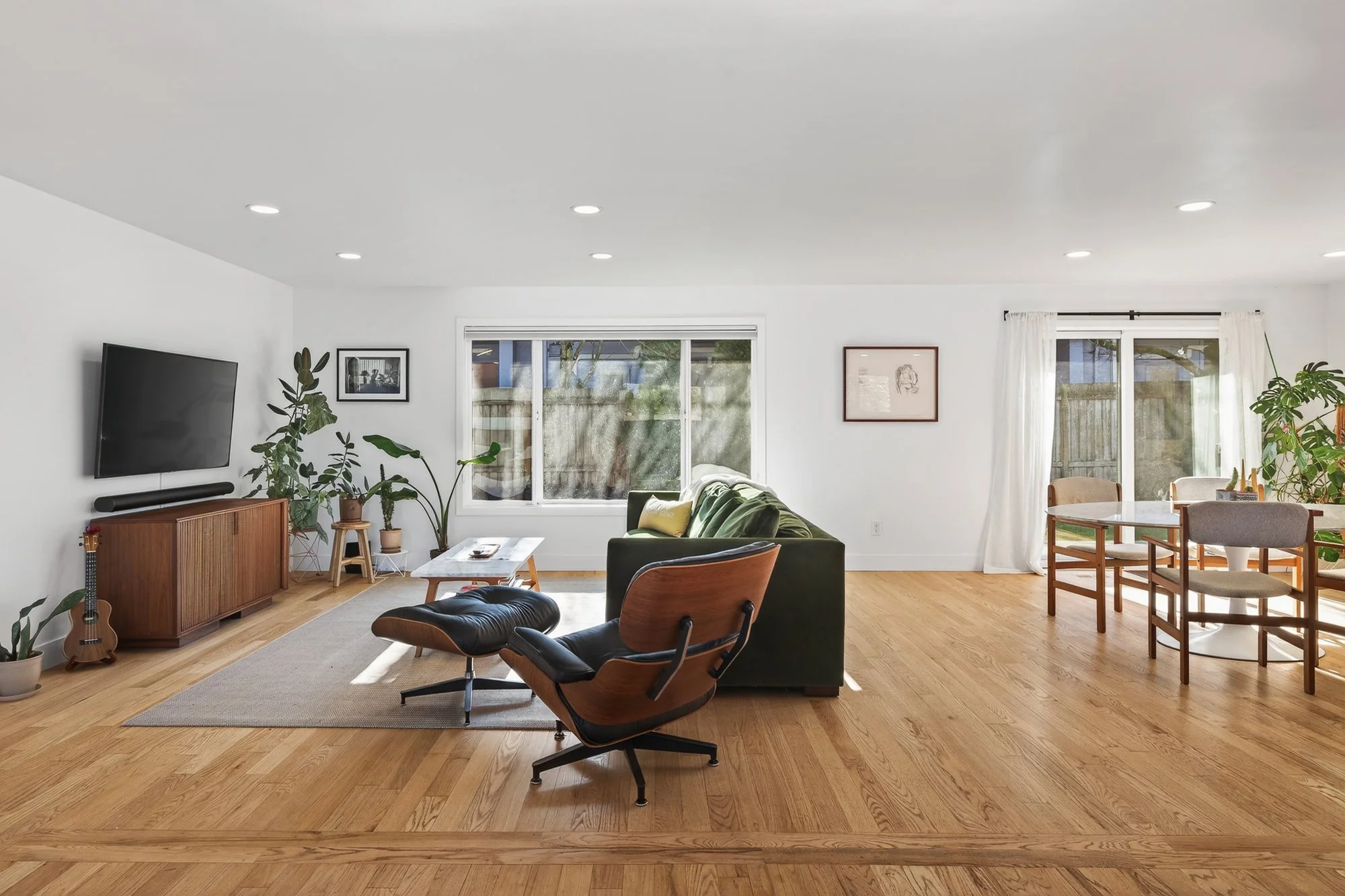 A spacious living room with hardwood flooring, white walls, and large windows. It features a green sofa, a wooden armchair with a black leather seat, a modern wooden media console with a flat-screen TV, and various potted plants. There is a small coffee table and framed artwork on the walls. A dining area with a round table and chairs is visible in the background, near a sliding glass door with white curtains.