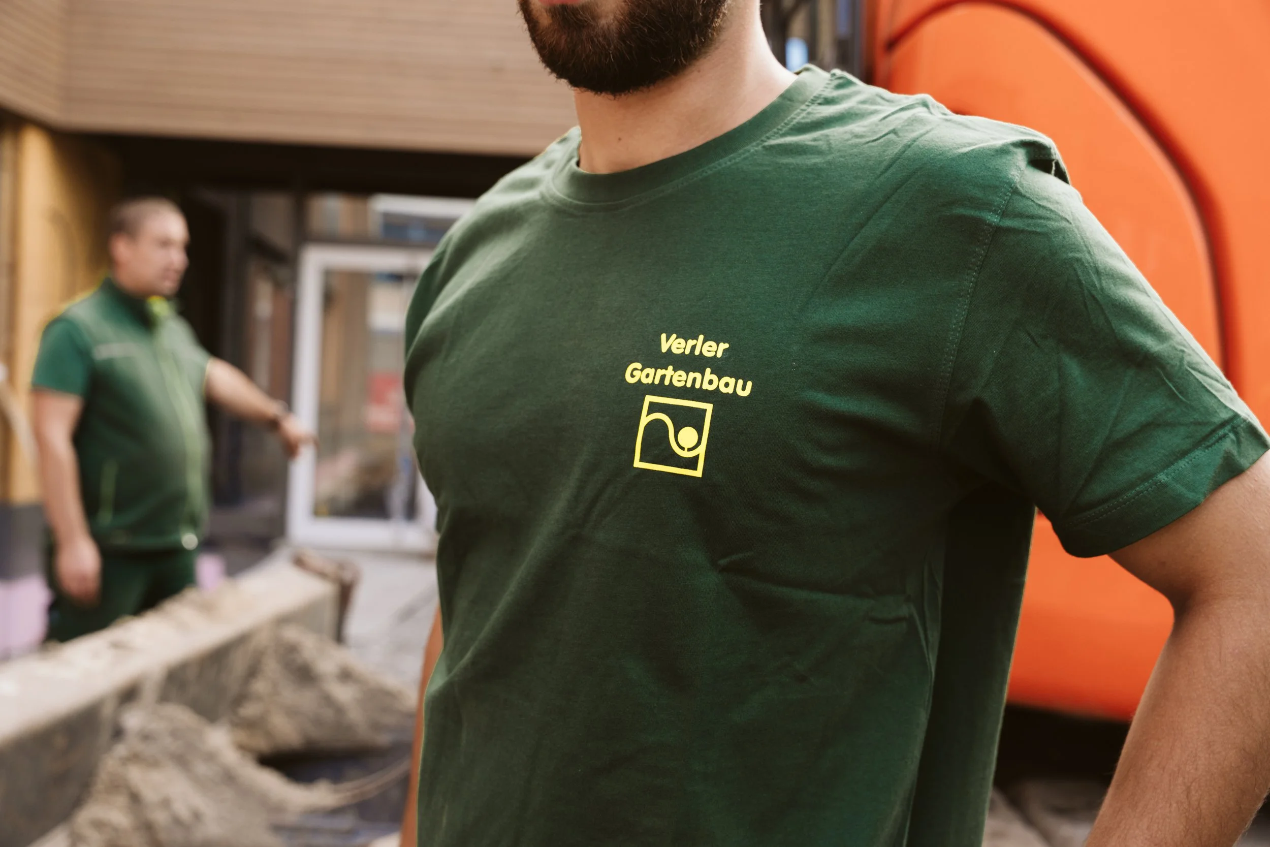 Close-up of a man wearing a green shirt with yellow text that reads "Verler Gartenbau" and a logo, standing indoors with another man in green in the background.