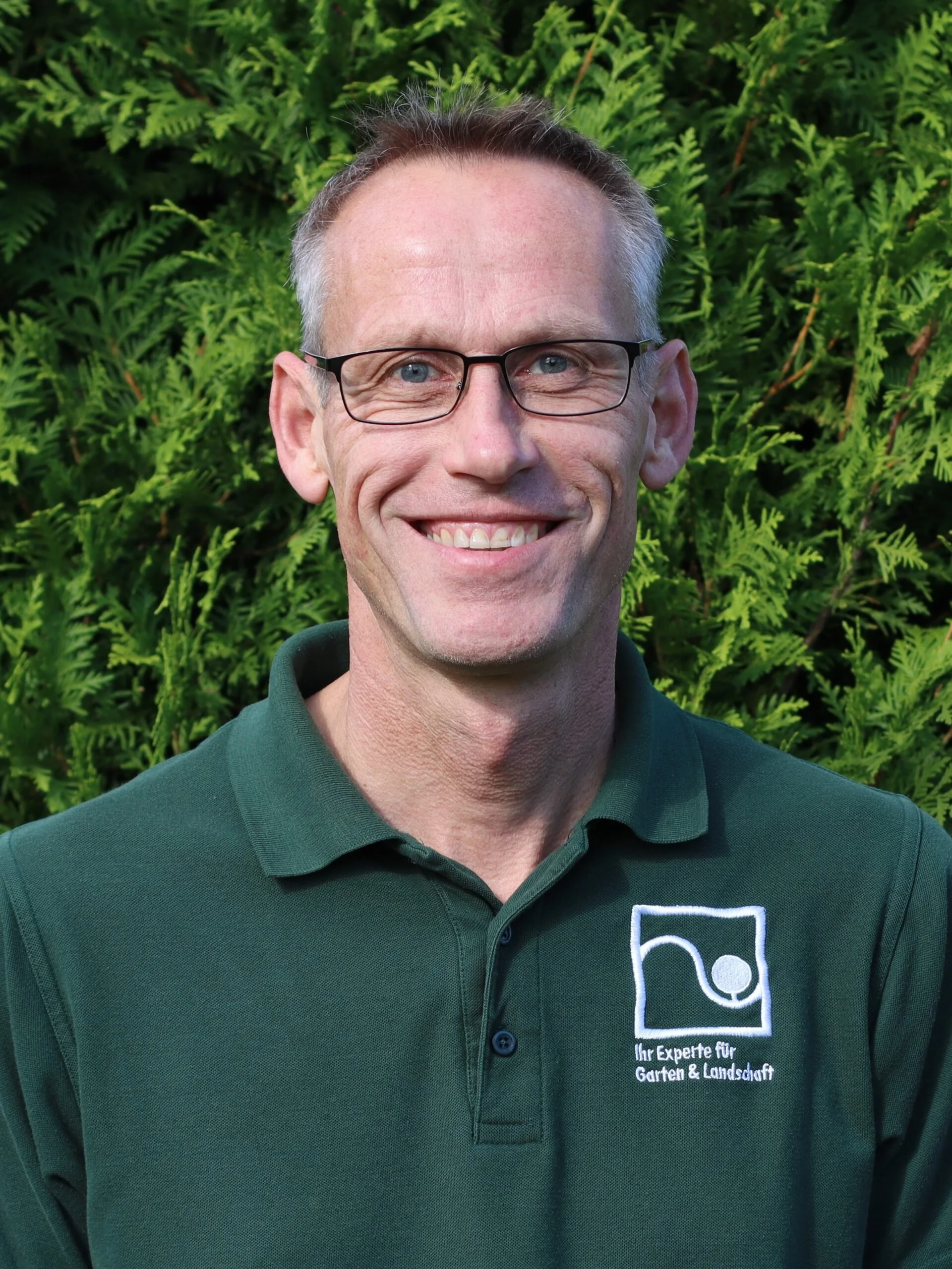 A man smiling, wearing glasses and a dark green polo shirt with a logo and German text, standing in front of green foliage.