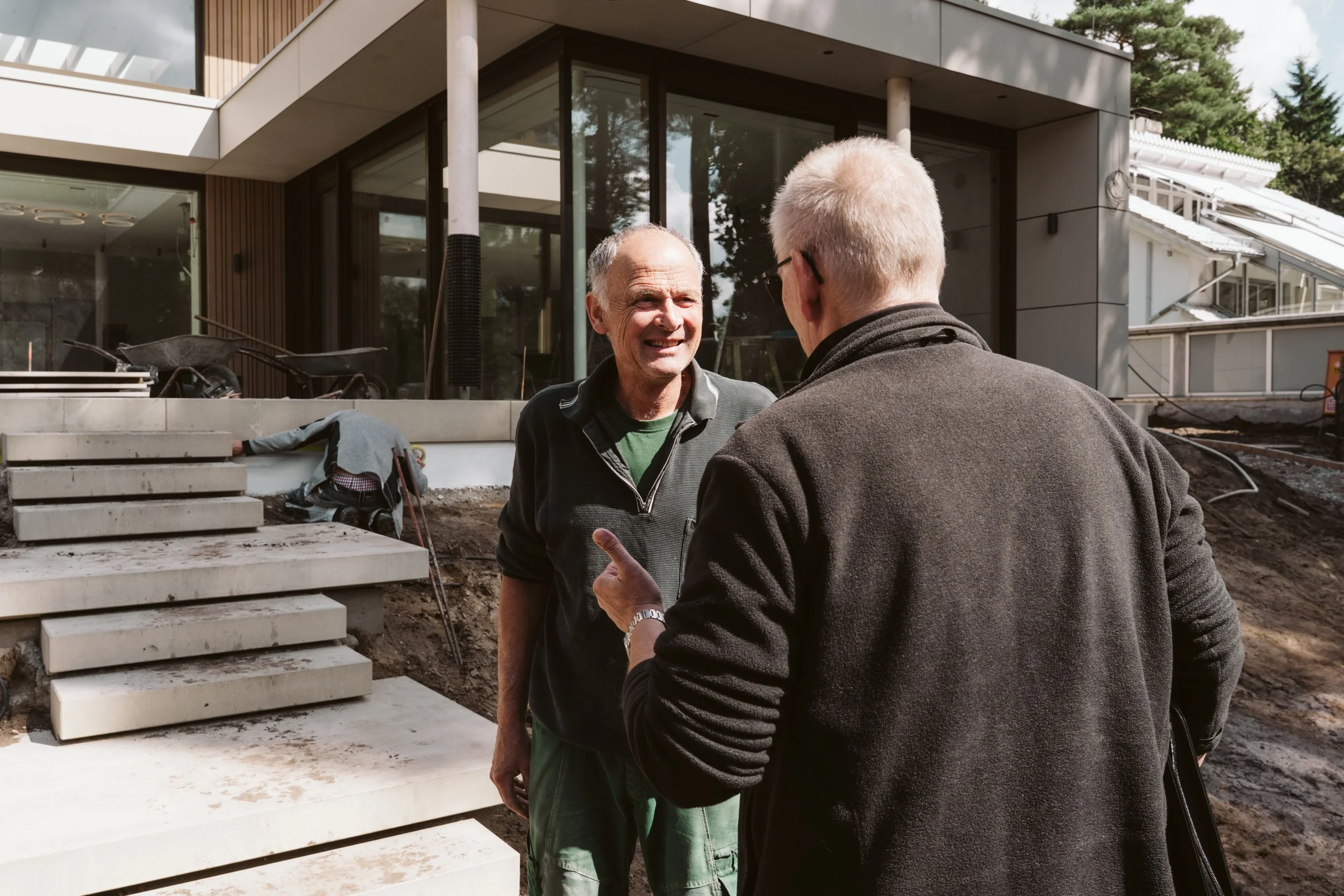 Two men engaged in conversation outdoors, one facing the camera smiling, in front of a modern building under construction.