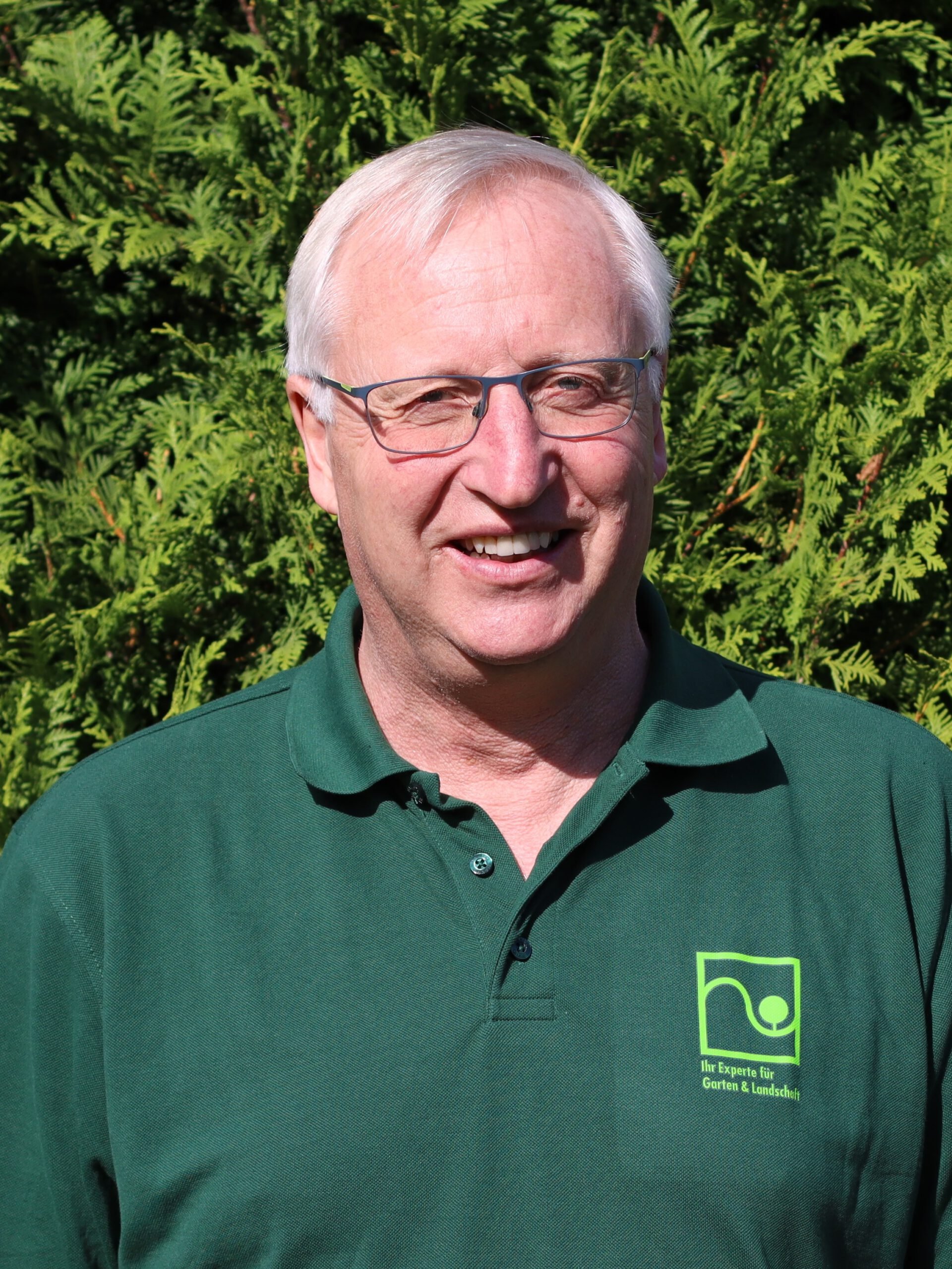 A smiling middle-aged man with glasses wearing a dark green polo shirt with a logo on the chest, standing outdoors in front of green leafy bushes.