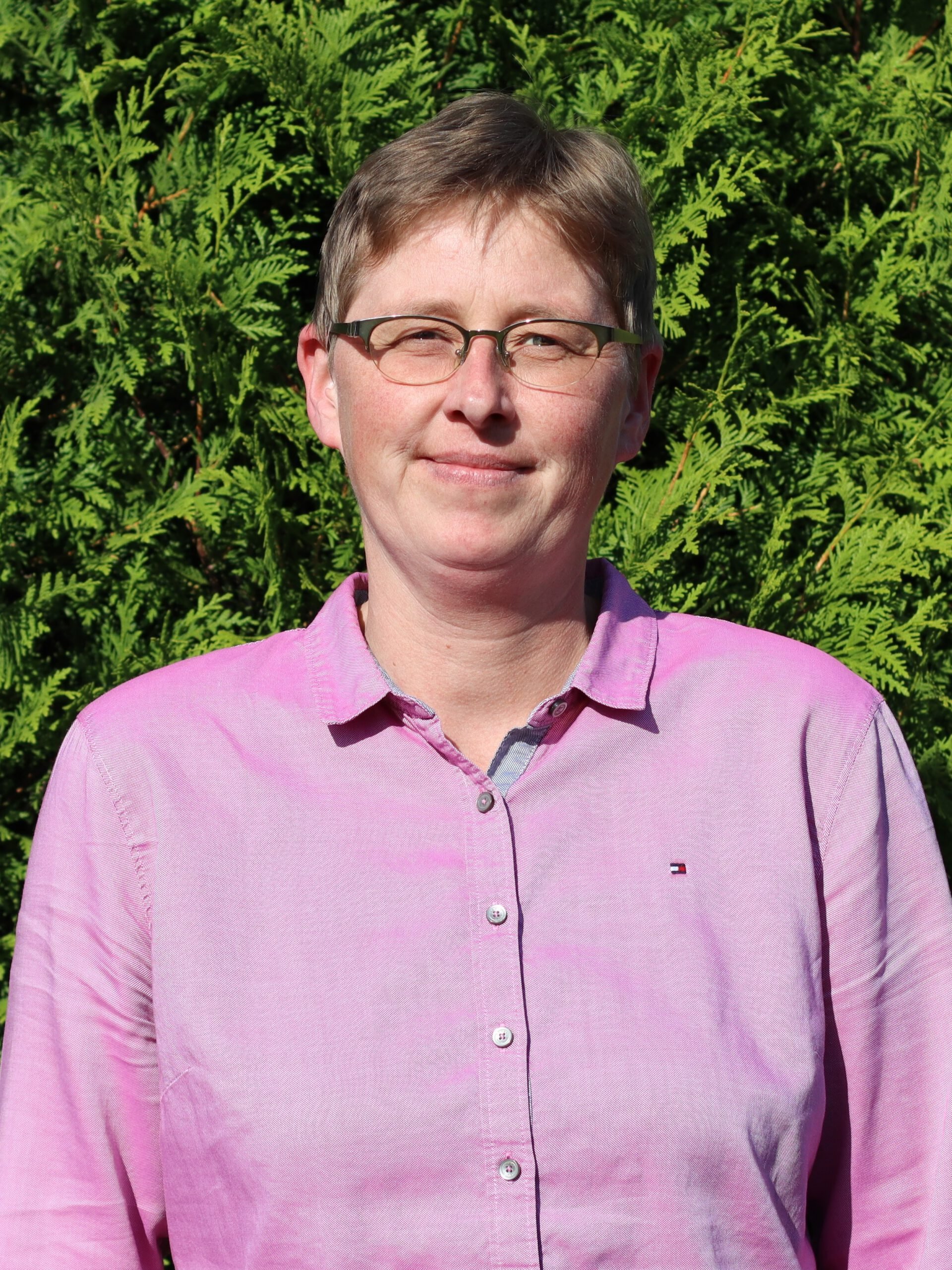 A woman with short brown hair and glasses, wearing a pink button-up shirt with a small logo, standing in front of green foliage.