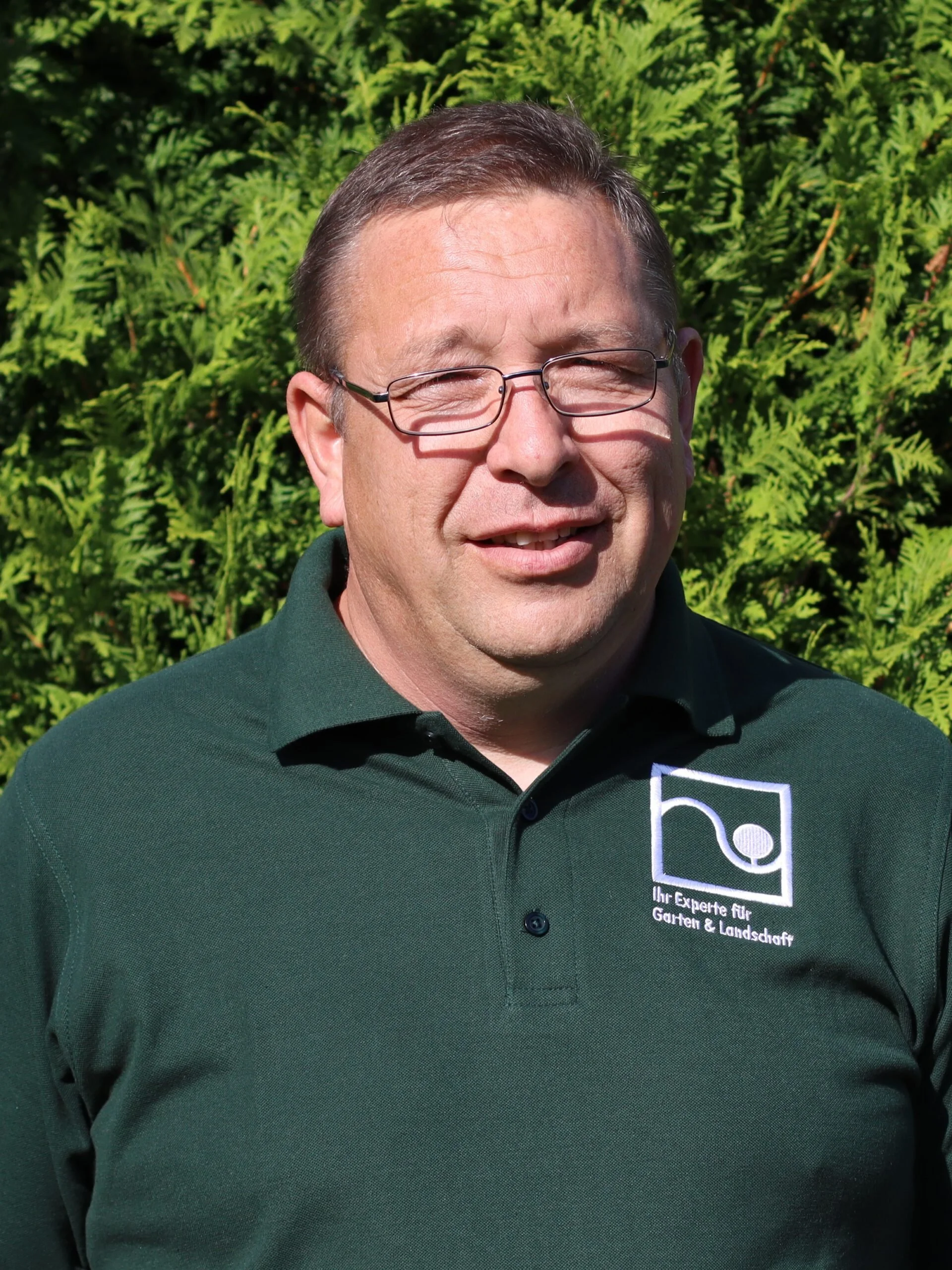 Man with glasses smiling outdoors, wearing a dark green polo shirt with a logo and text that says 'Ihr Experte für Garten & Landschaft' on it, standing in front of green foliage.