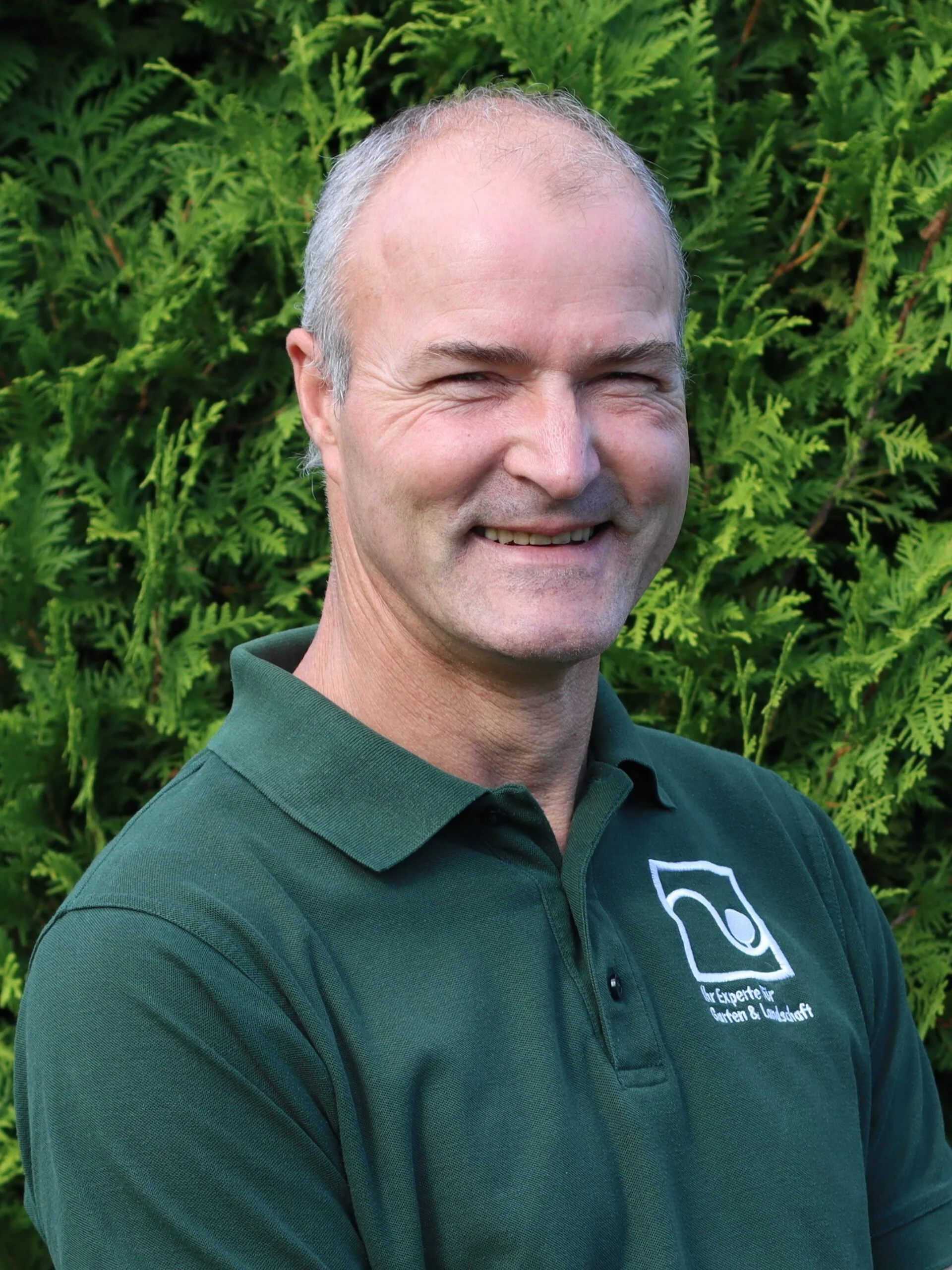 A smiling middle-aged man with short gray hair, wearing a dark green collared shirt with a logo and text, standing outdoors in front of green foliage.