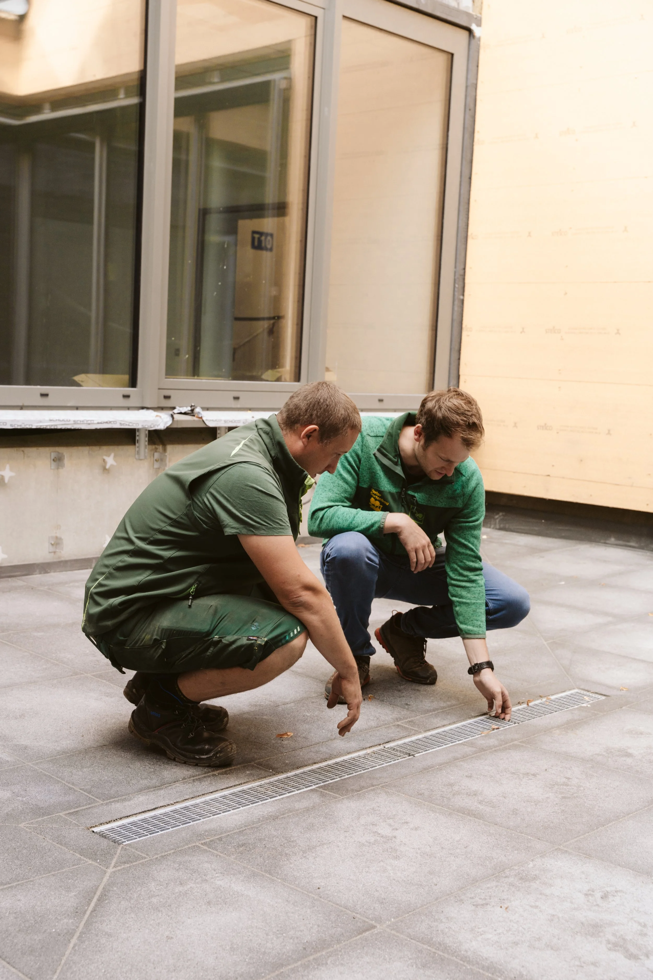 Two men crouching down and looking at the drain on the floor of a construction site or unfinished building.