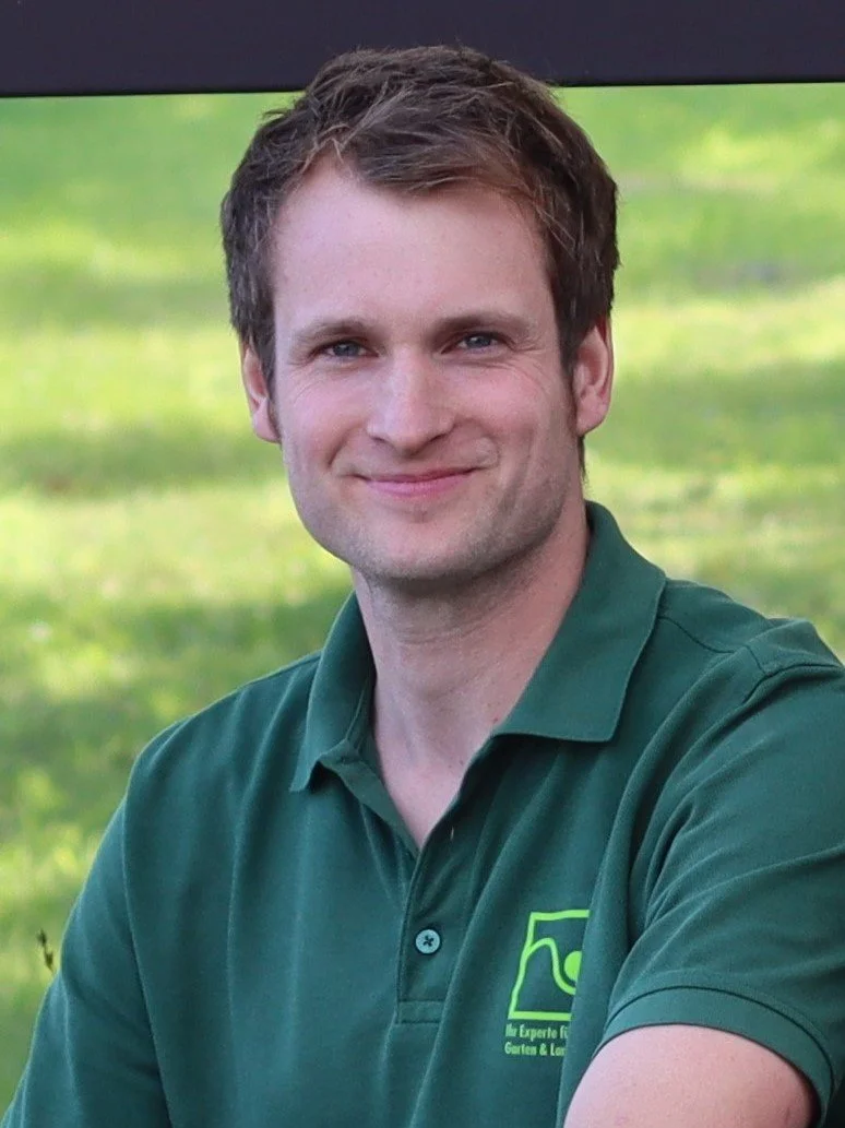 A young man with short brown hair, fair skin, and blue eyes smiling, wearing a dark green polo shirt with a logo on it, standing outdoors with a blurred background of green grass.