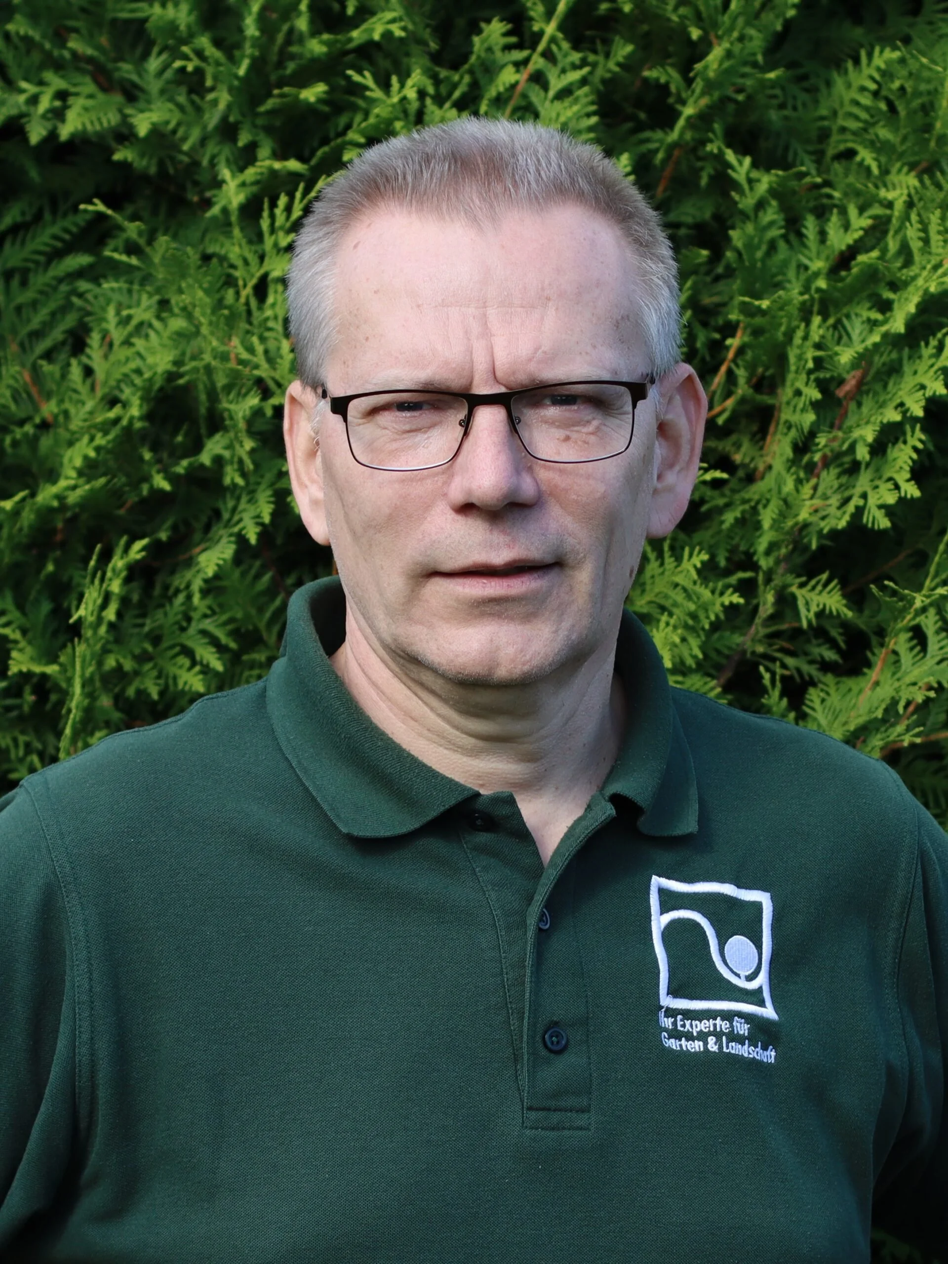 Close-up of man wearing glasses and a dark green collared shirt with logo, standing in front of green foliage.