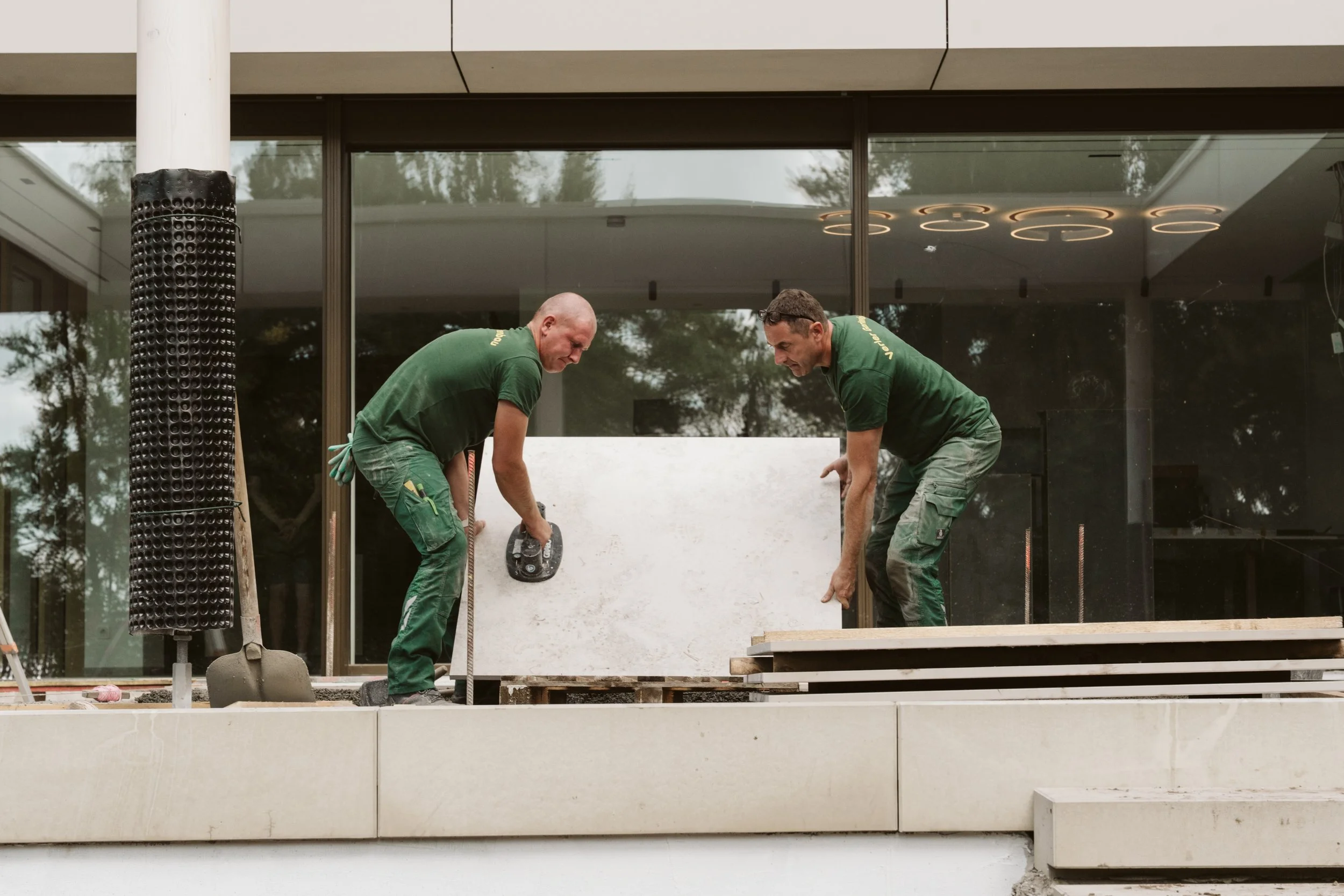 Two construction workers are installing a white stone or concrete panel on an exterior building facade. They are focused on their work, with one holding a tool and the other adjusting the panel. The building has a large glass window or sliding door in the background reflected with trees and sky, and there is a cylindrical black ventilation or exhaust pipe on the left side of the image.
