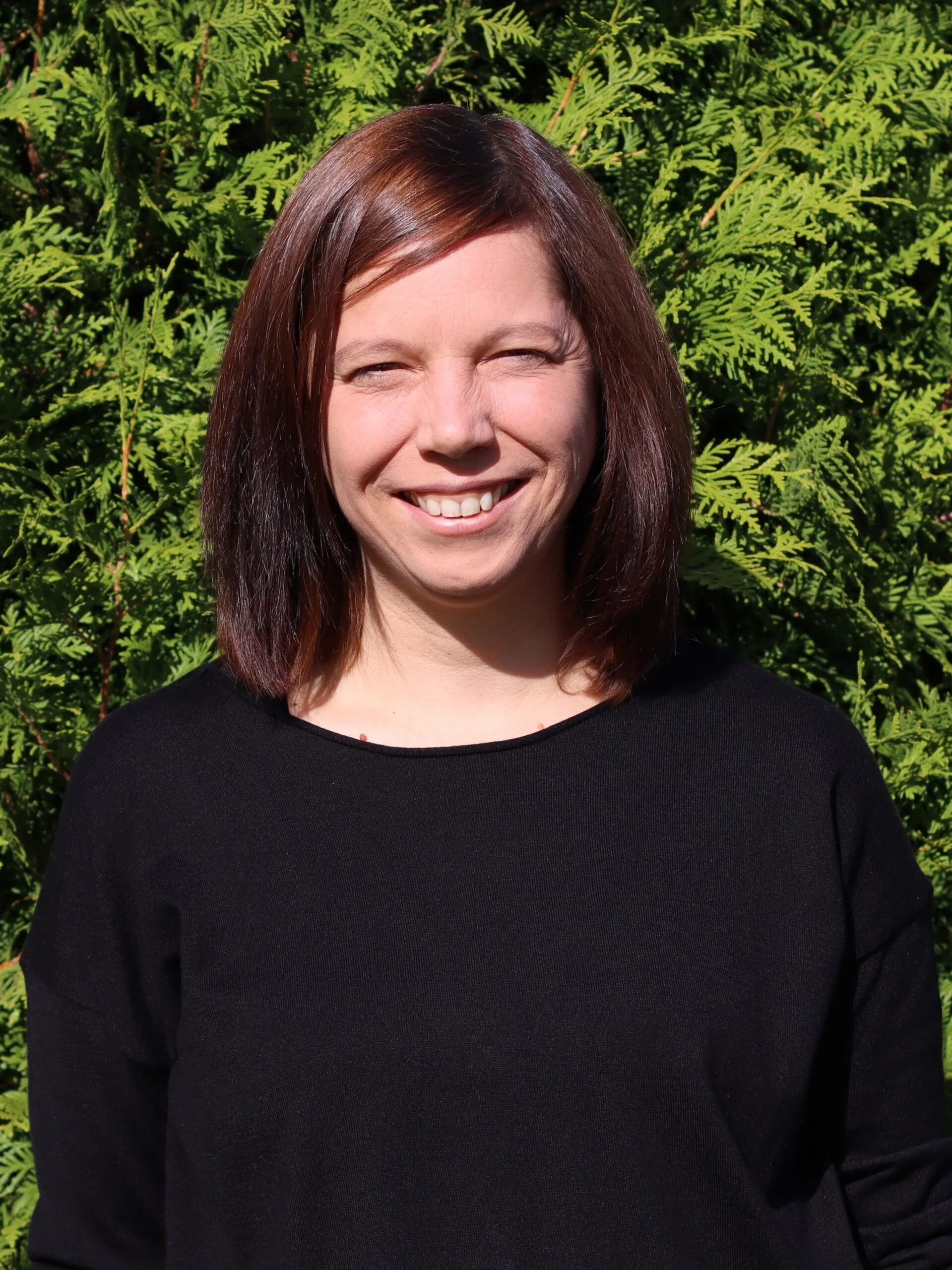 A woman with shoulder-length brown hair smiling in front of lush green ferns.