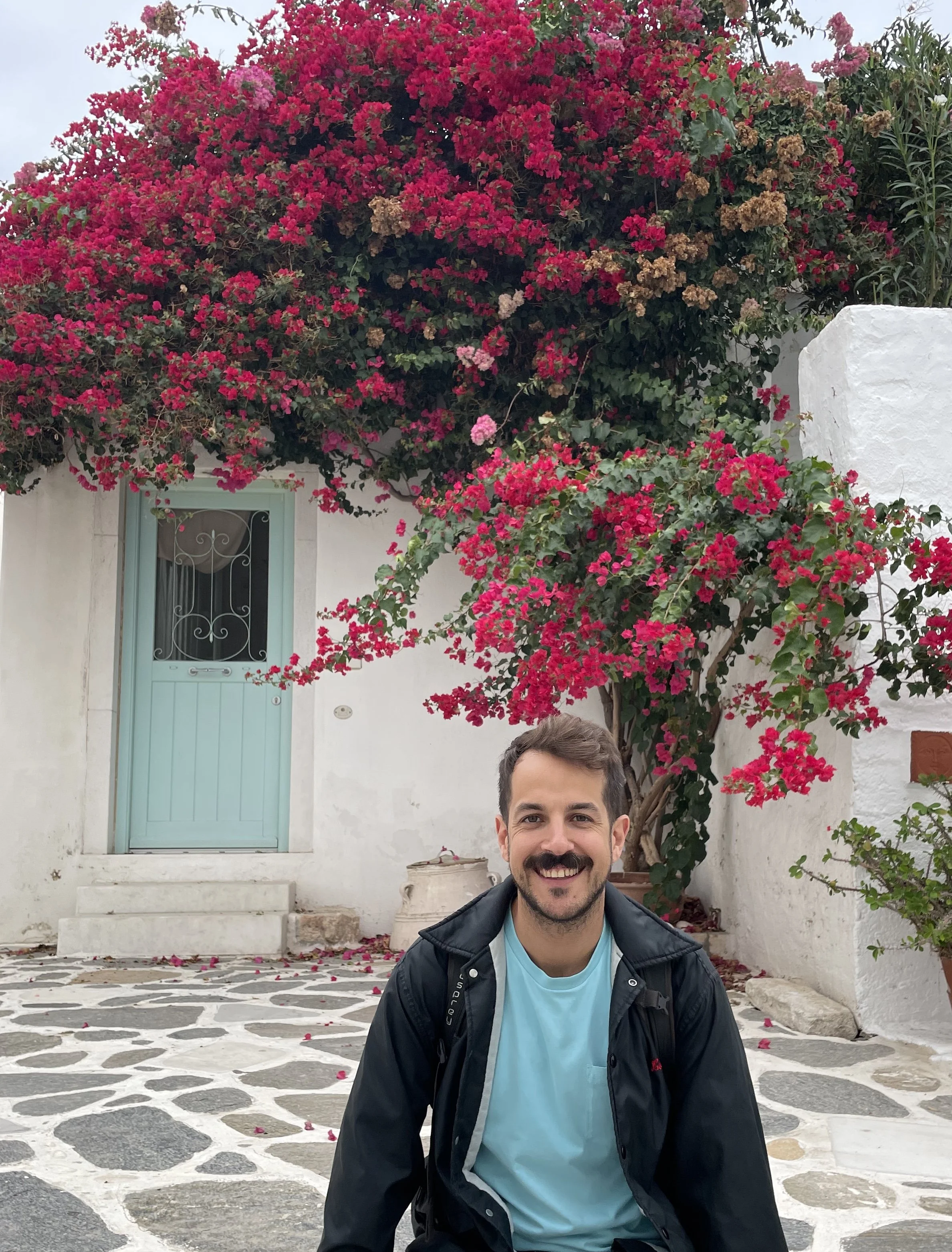 Hombre sonriendo en un patio con puerta pintada de azul y plantas de buganvilla de flores rosadas y moradas.