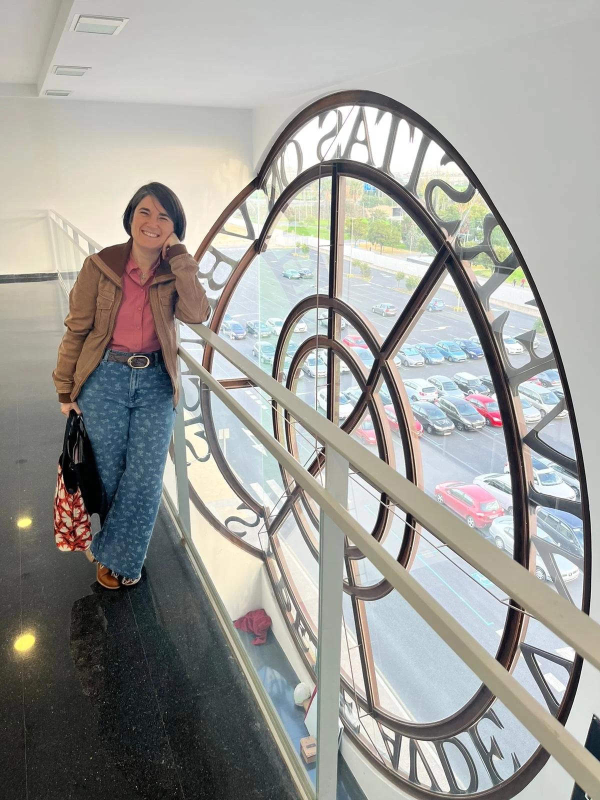 Mujer sonriendo en interior cerca de una gran ventana con forma de reloj, estacionamiento visible afuera.