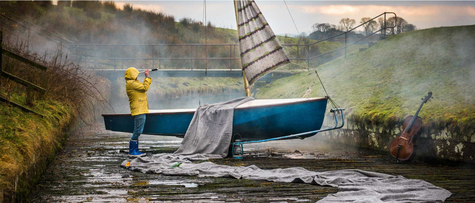 A person in a yellow raincoat and blue rain boots standing on a dock, pointing a telescope towards the sky. A small blue sailboat is on the dock with a grey blanket draped over the side. A cello leans against the grassy bank on the right, and a lantern is on the dock near the boat. The background shows a foggy, hilly landscape with trees and a wooden bridge.