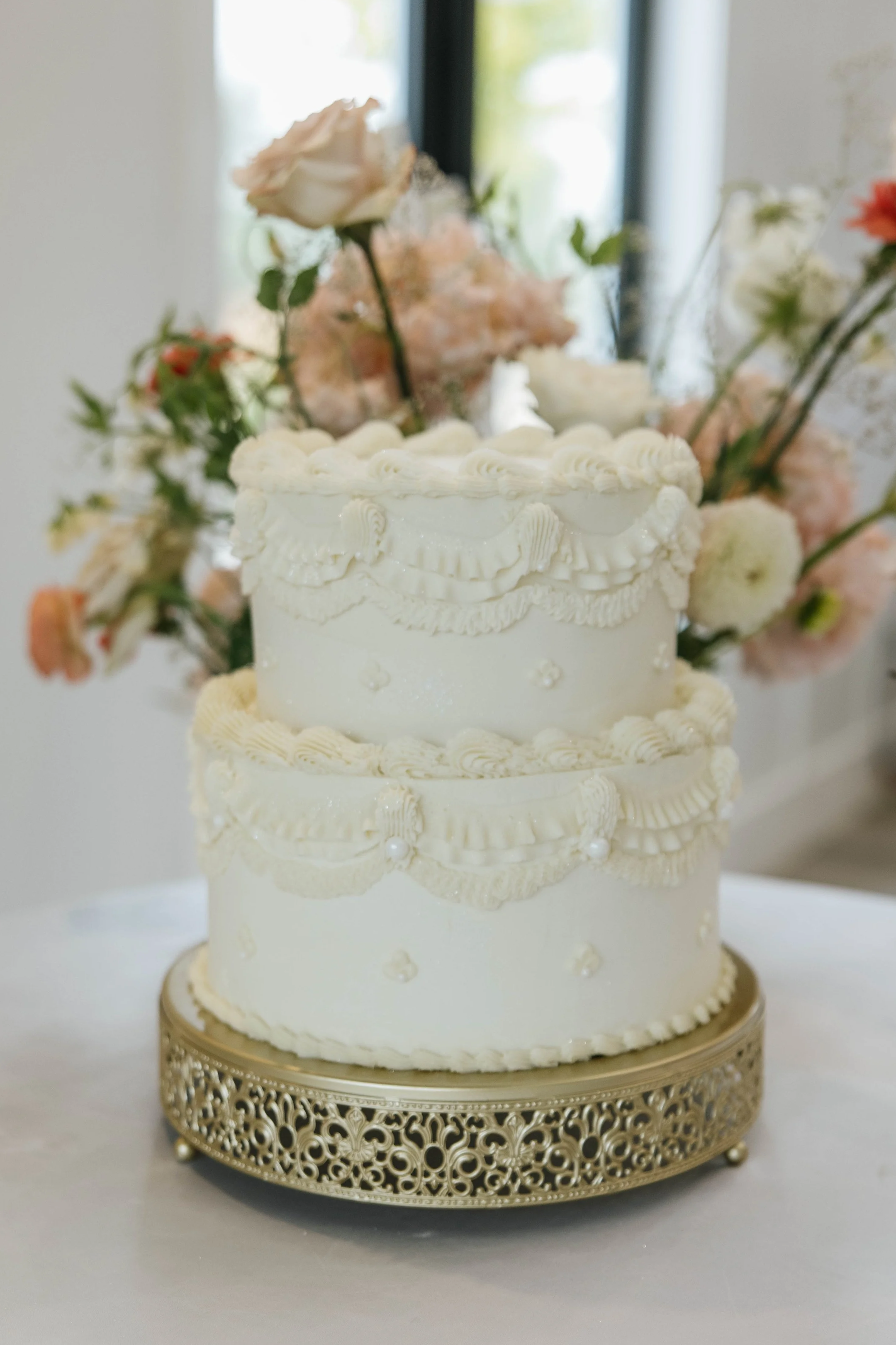 A three-tiered white wedding cake with intricate piping decorations, placed on a decorative gold cake stand, with a bouquet of pink and white flowers in the background.
