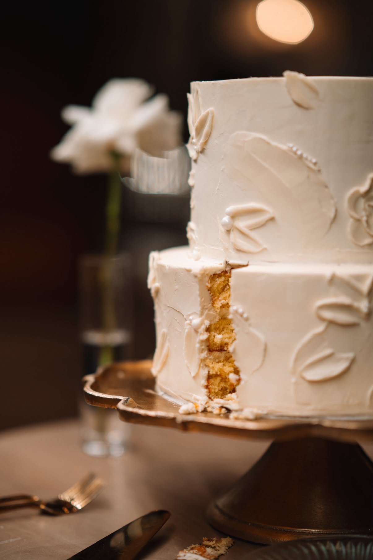 A two-tiered white cake with decorative icing, partially cut to reveal the inside, placed on a cake stand with a cake server nearby, and a white flower in a small vase in the blurred background.