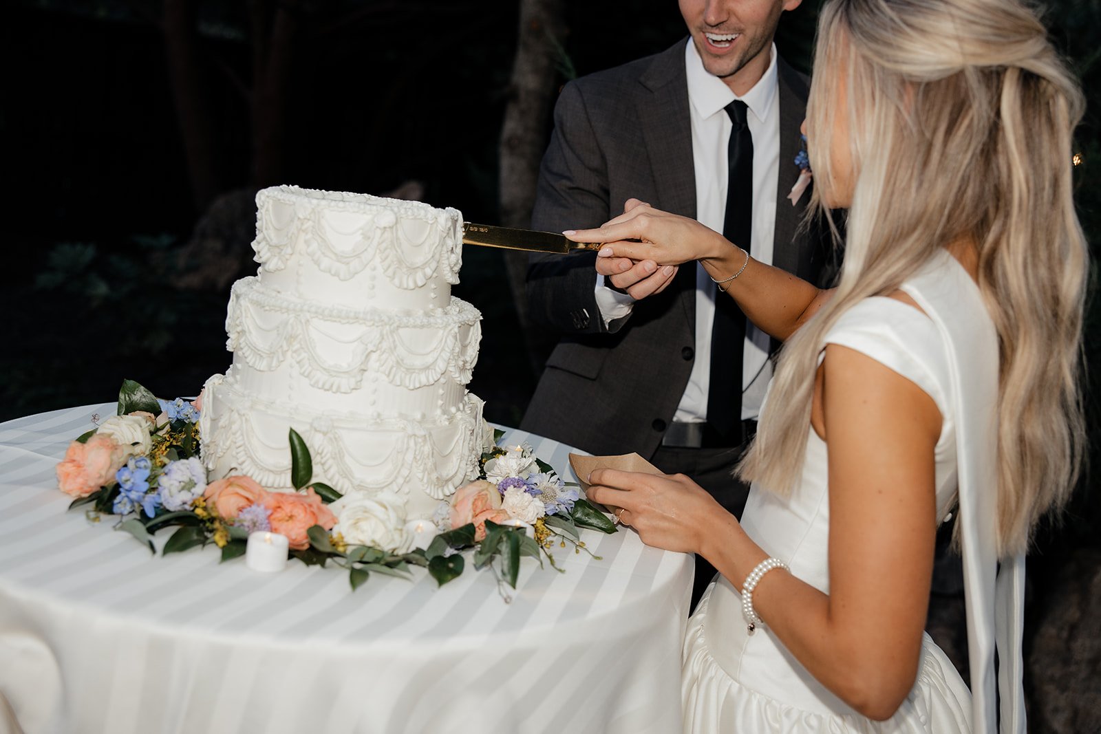 A bride and groom cutting a wedding cake decorated with white icing and floral arrangements during their wedding celebration.