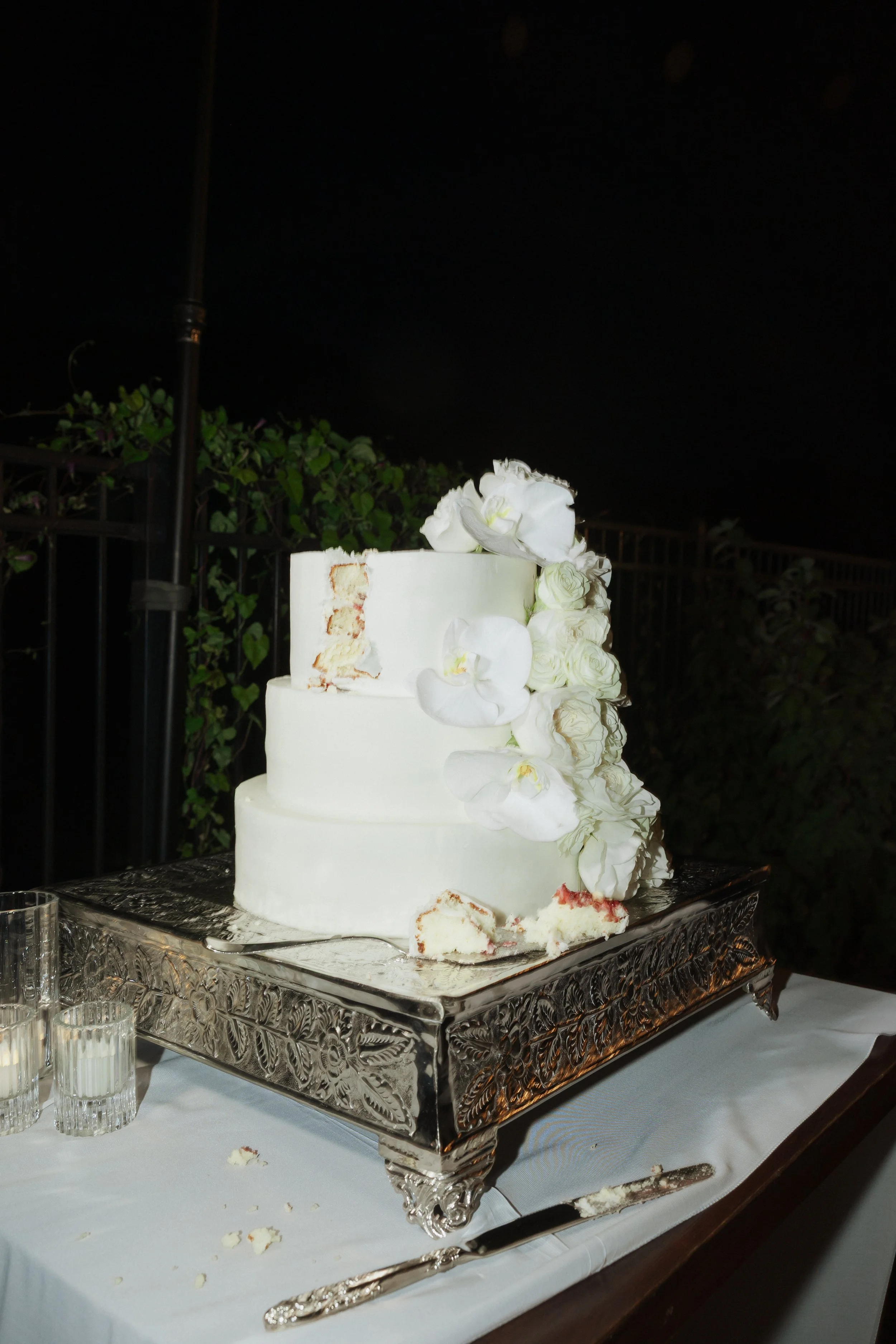A three-tiered white wedding cake decorated with white flowers on top and cascading down the side, placed on a decorative silver cake stand.