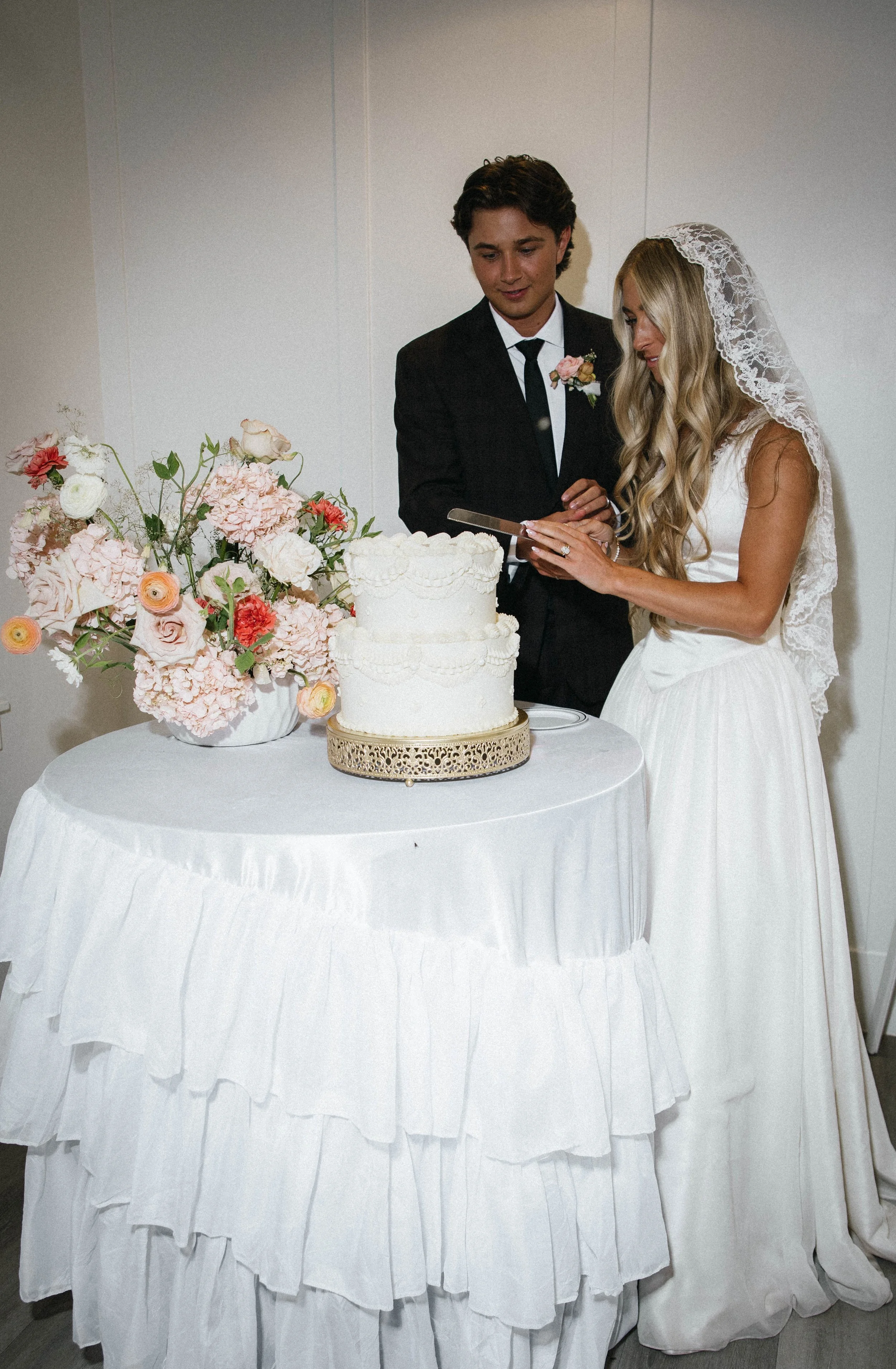 A bride and groom standing behind a wedding cake and floral arrangement, looking at a tablet together at their wedding reception.