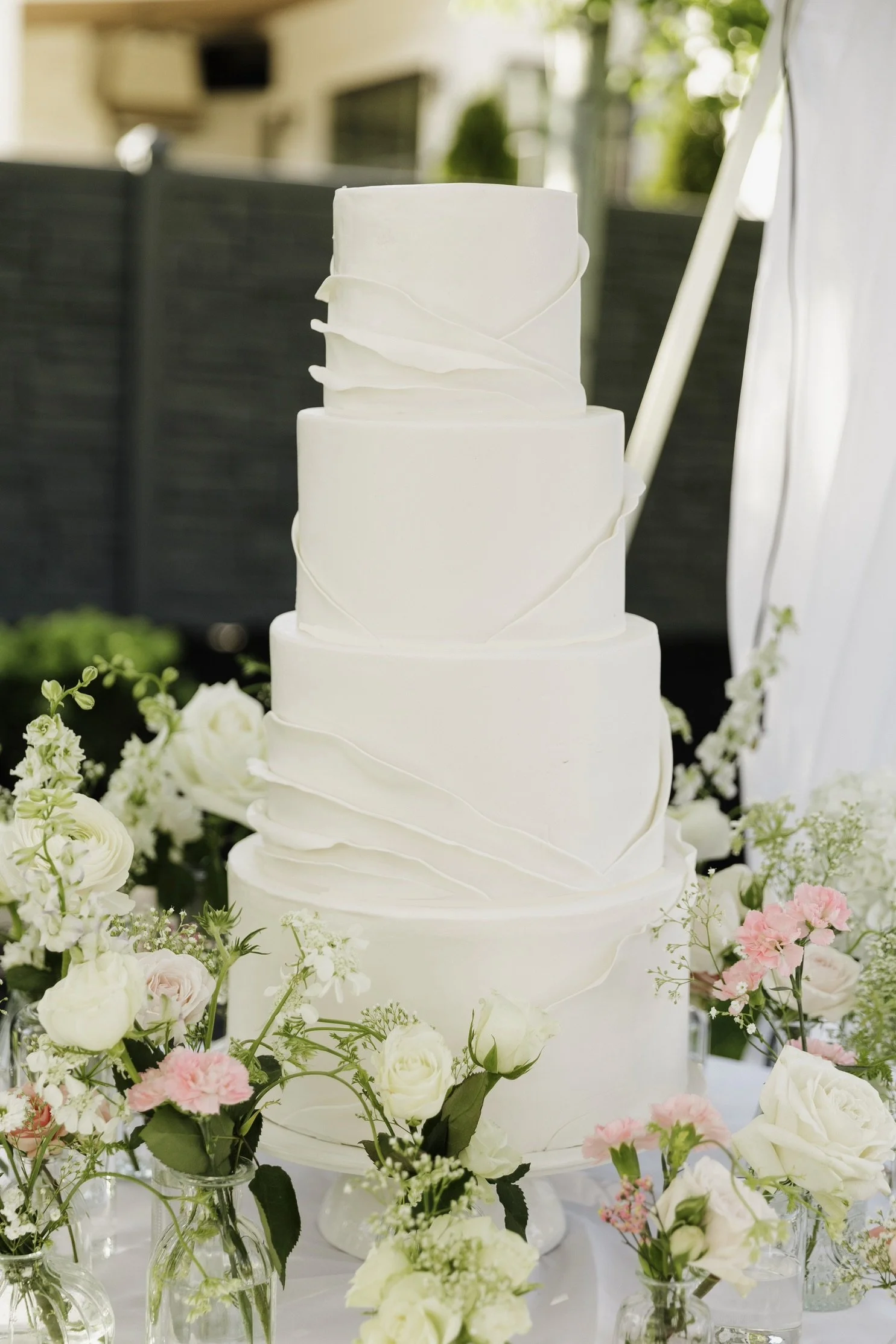 A white, tiered wedding cake decorated with elegant white fondant ribbon-like accents, surrounded by an array of white and pink flowers in glass vases, set outdoors with sunlight.