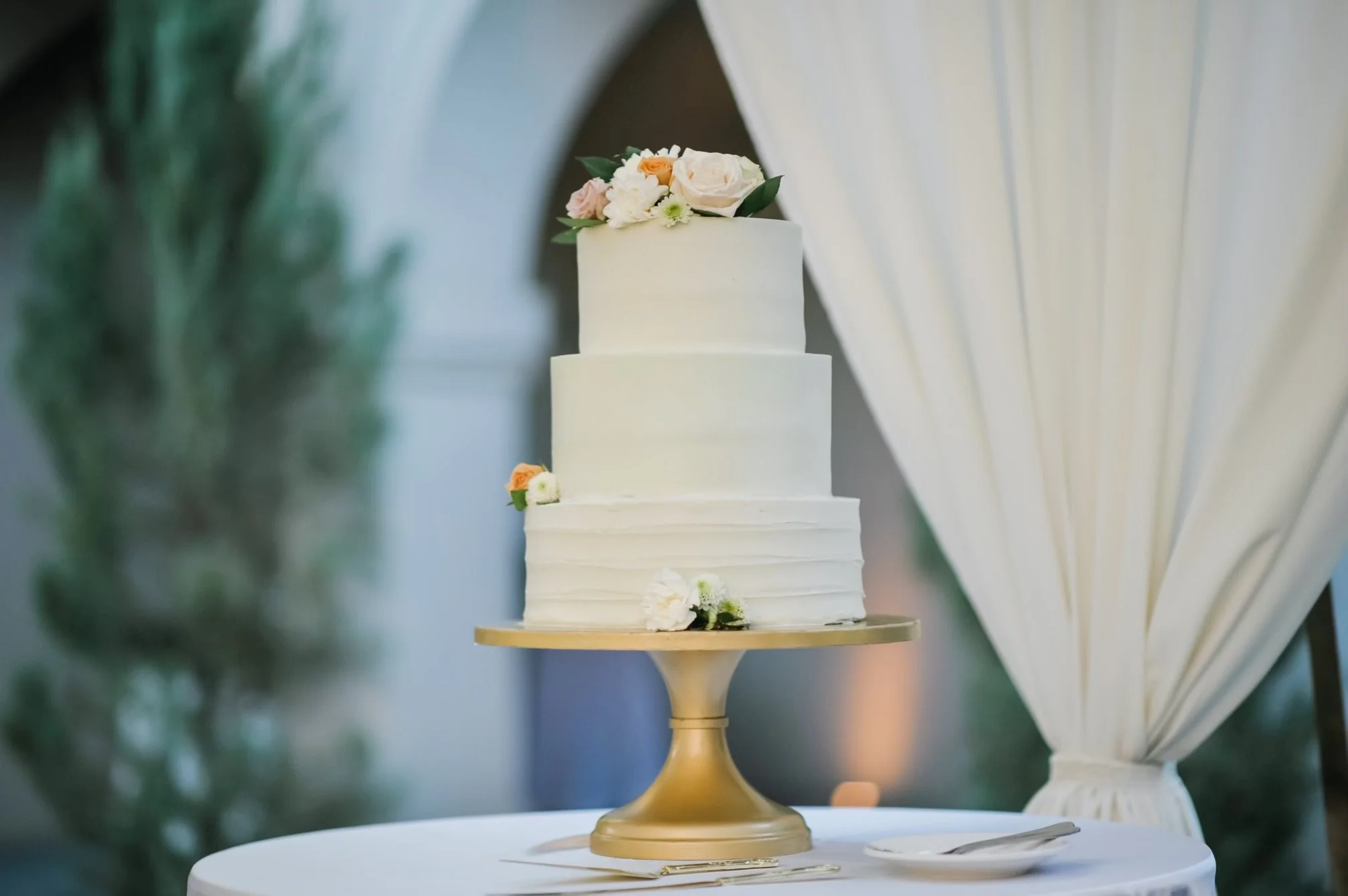 A white, two-tiered wedding cake decorated with pastel-colored flowers, displayed on a gold cake stand at a wedding reception.