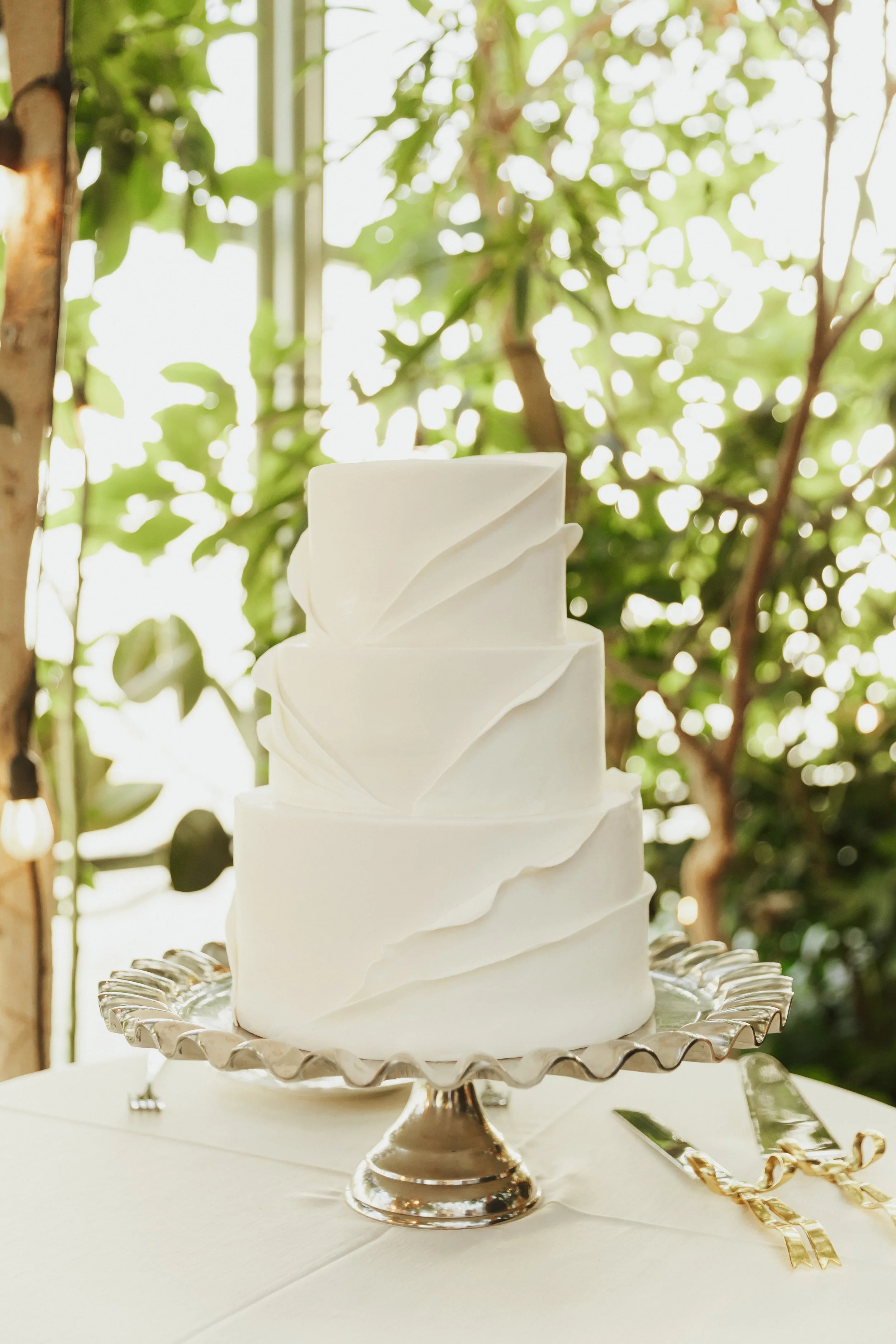 A multi-tiered white wedding cake with elegant ribbon-like decorations on each tier, displayed on a silver cake stand with a scalloped edge, set against a backdrop of green leaves and bright sunlight.