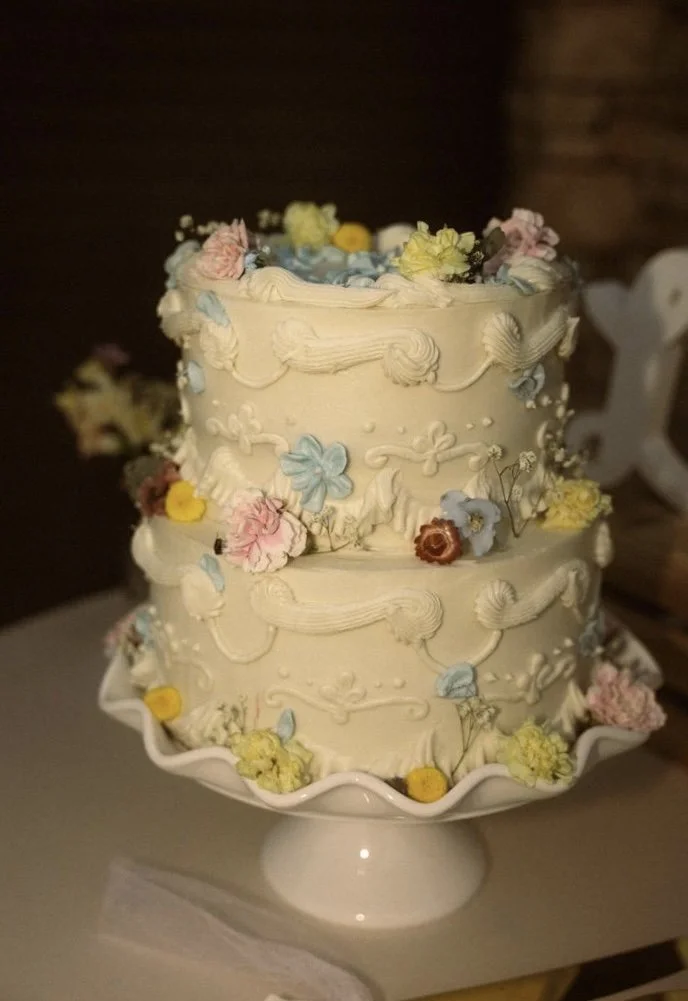 A two-tier cream-colored wedding cake decorated with pastel-colored flowers and piped icing designs, placed on a white cake stand.