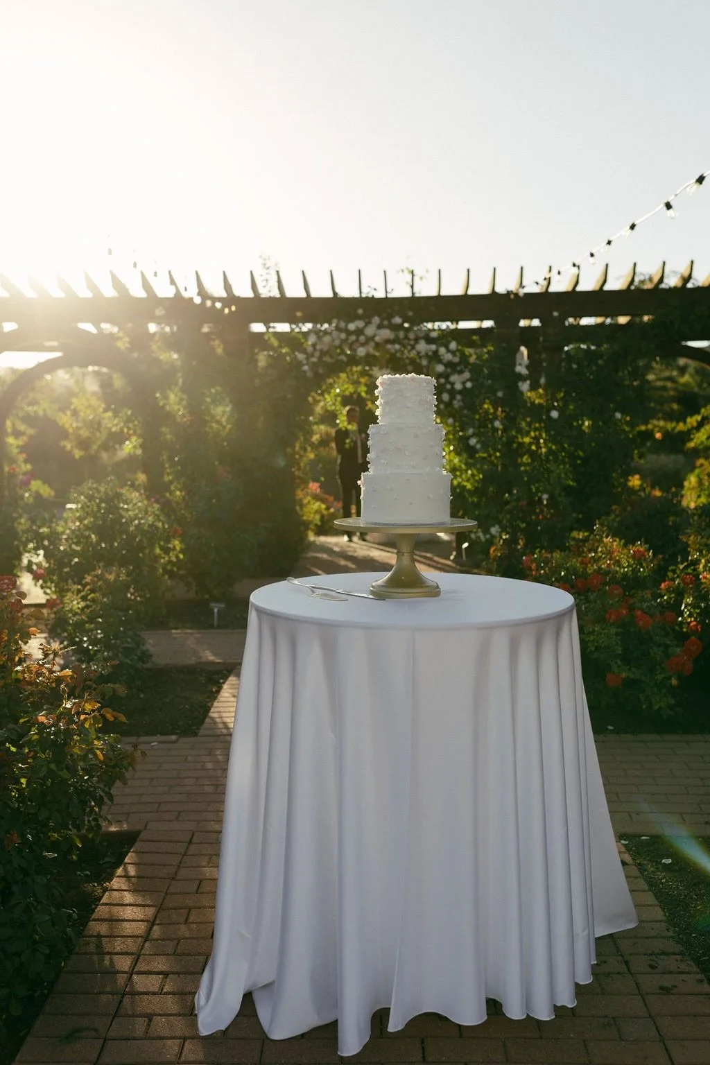 A white three-tiered cake on a cake stand, placed on a round table with a white tablecloth, outdoors during sunset with flowers and greenery in the background.
