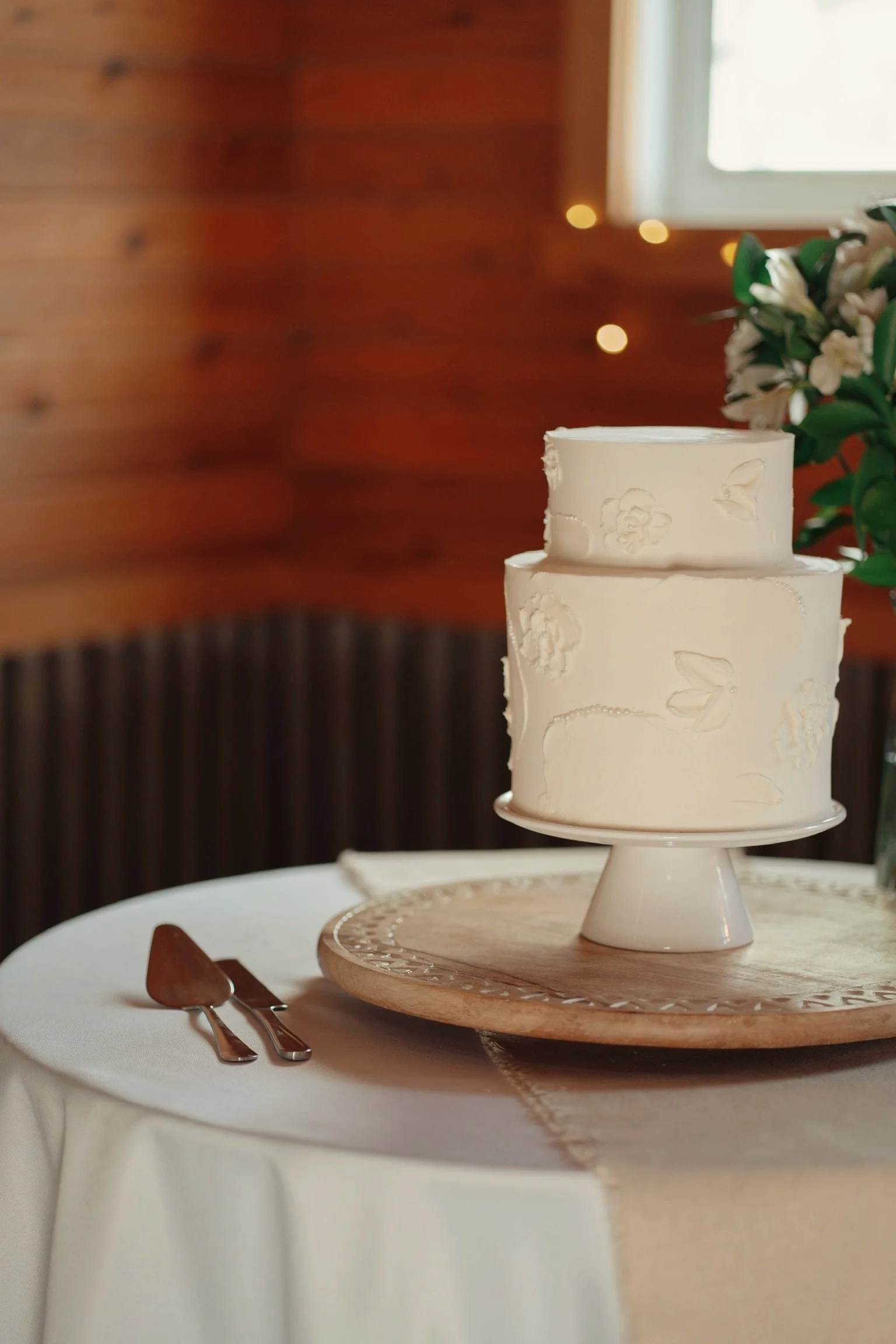 A three-tiered white wedding cake with flower and bird designs on a cake stand, placed on a wooden round tray, on a table with a white tablecloth, next to a small fork, and with a blurred background of a wooden wall and window with curtains.