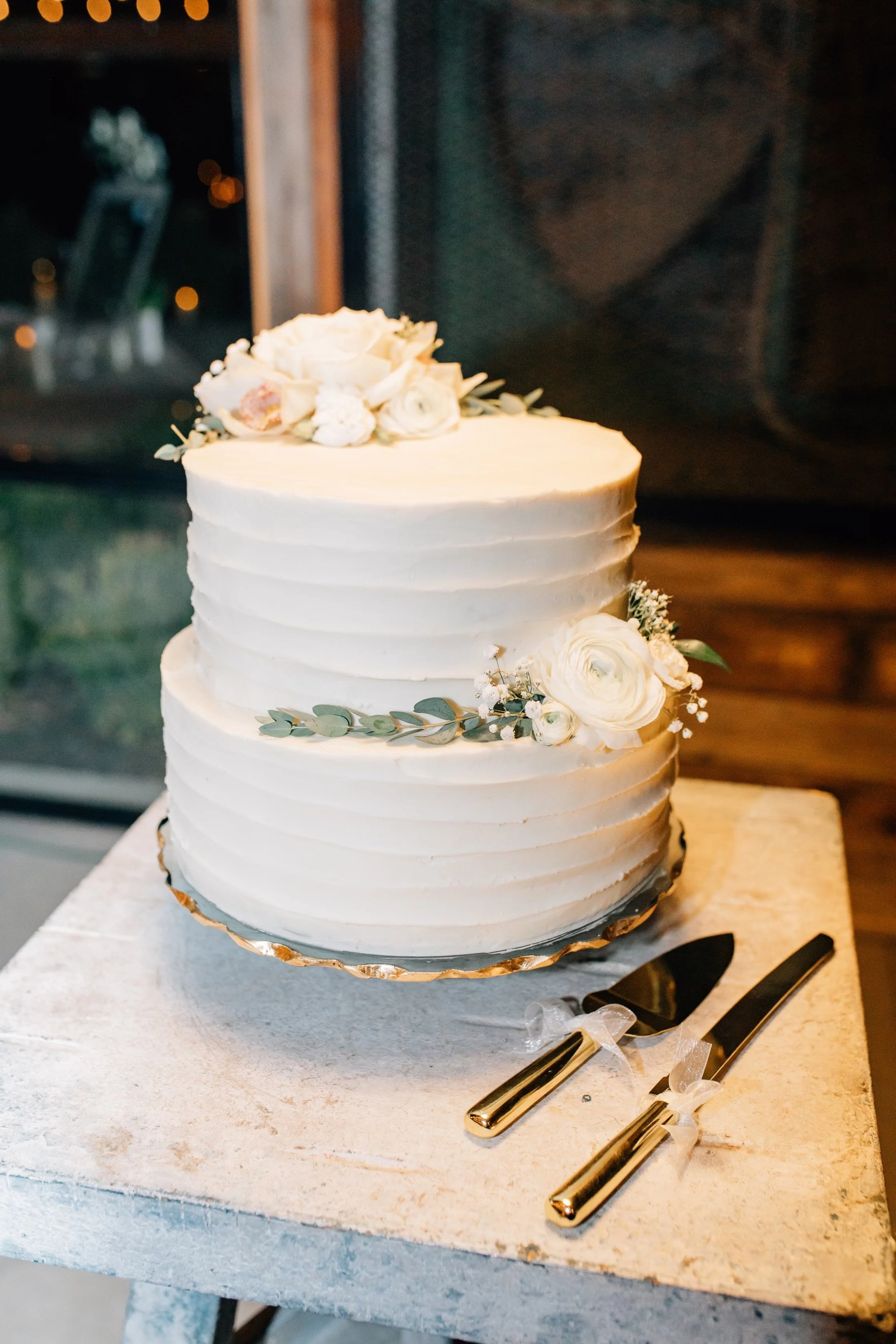 A white wedding cake with white flowers and green leaves on top, placed on a white table with gold-colored cake knife and server beside it.