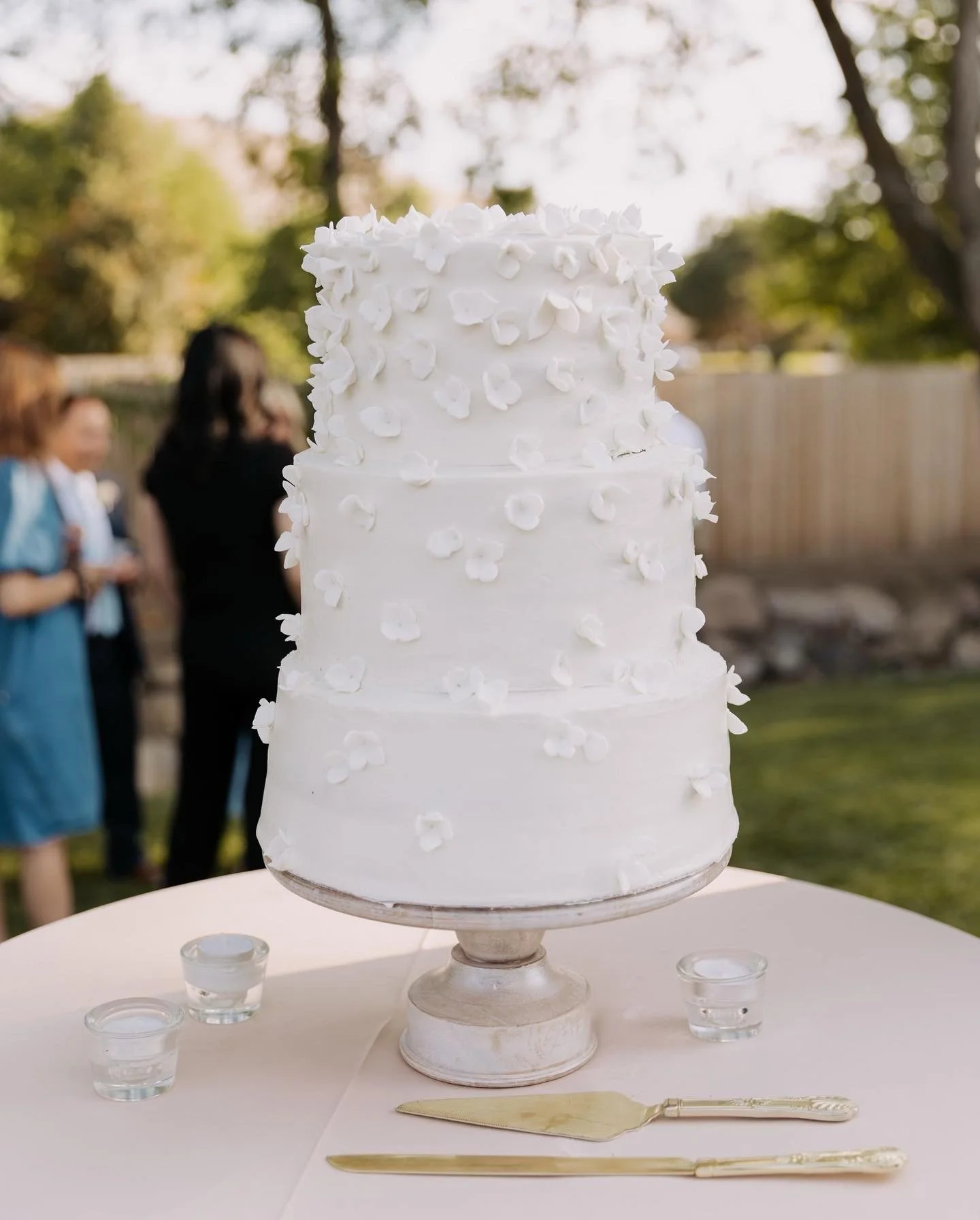 A tall white wedding cake with white floral decorations, placed on a glass cake stand on a white table. In the background, people at an outdoor event can be seen, with trees and a fence.