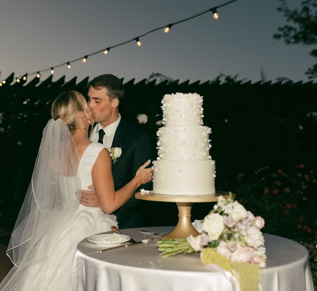 A bride and groom kiss during their wedding reception outdoors at night, with a three-tiered white wedding cake and a floral bouquet on the table in front of them, as string lights hang above.